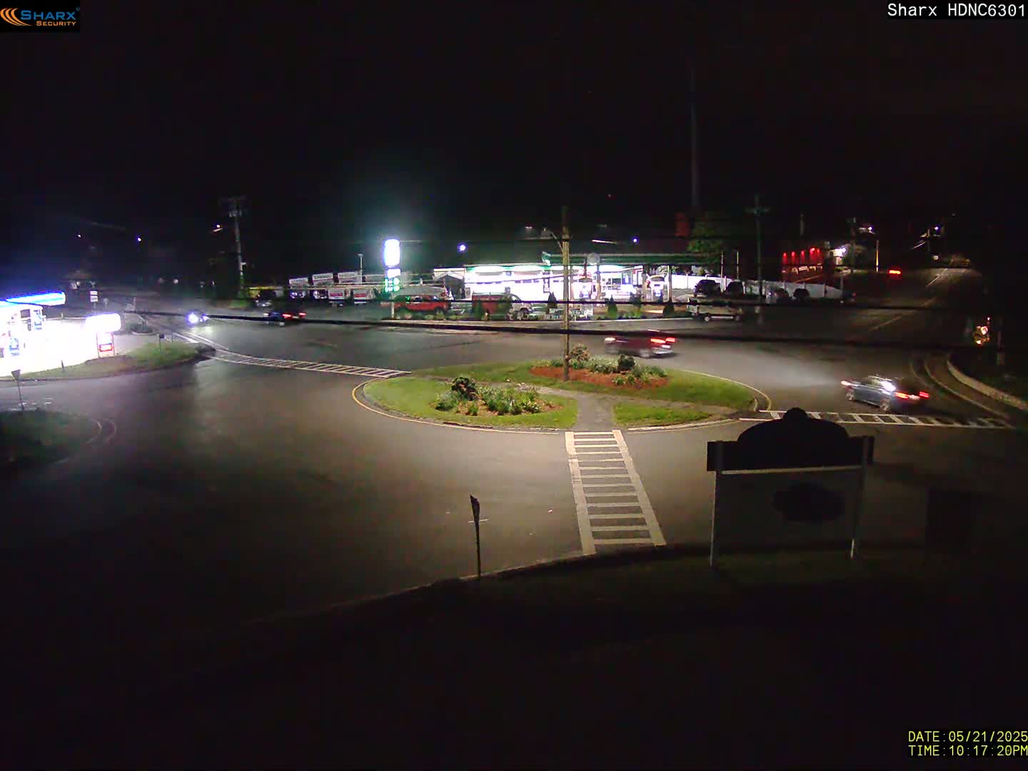 A nighttime view of a roundabout with several vehicles and a gas station in the background.