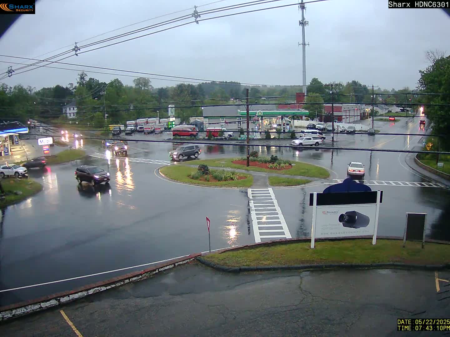 A rain-soaked roundabout with several cars and a gas station is visible during an overcast day.