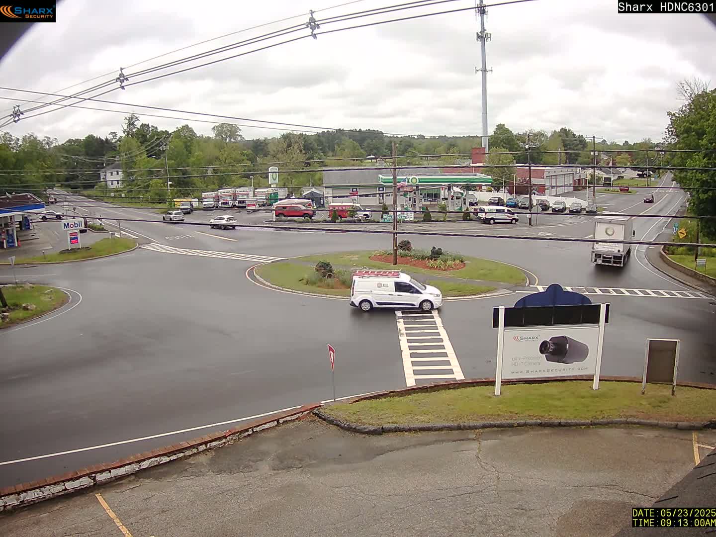 A wet roundabout and roads with several vehicles, including a white van, are surrounded by buildings, gas stations, and trees under an overcast sky.