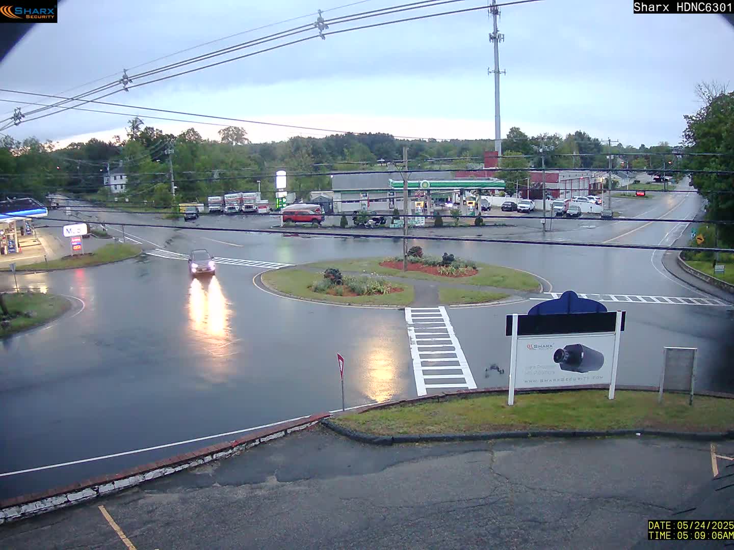 A wet, overcast day at a roundabout intersection with a gas station and several vehicles visible.