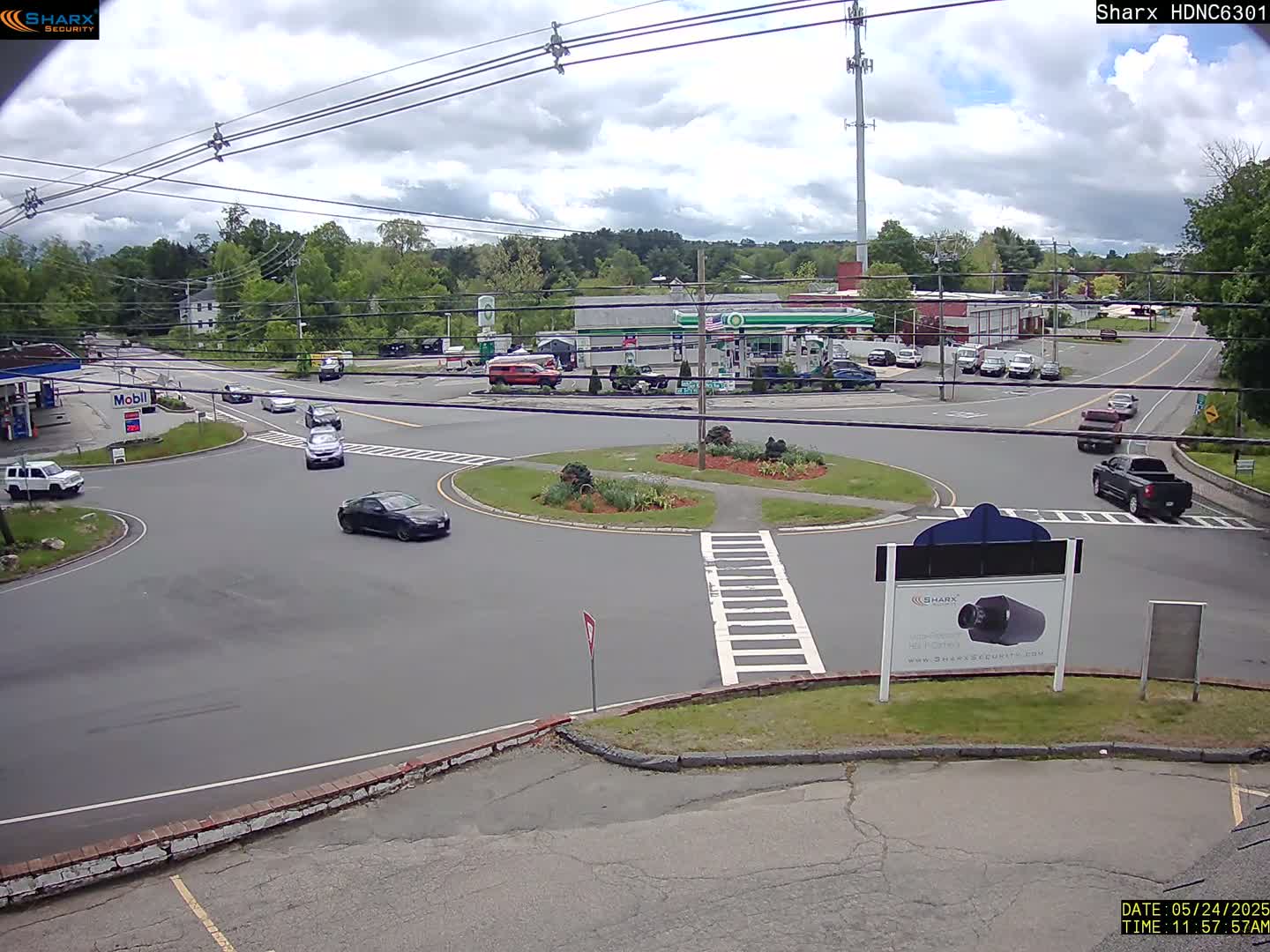 A mostly sunny day shows several cars navigating a roundabout near a gas station and other businesses, with a few cars parked along the side of the road.