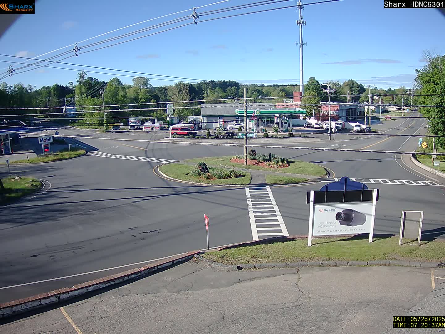 A sunny day reveals a road intersection with a traffic circle, gas station, and other businesses, surrounded by trees and greenery.