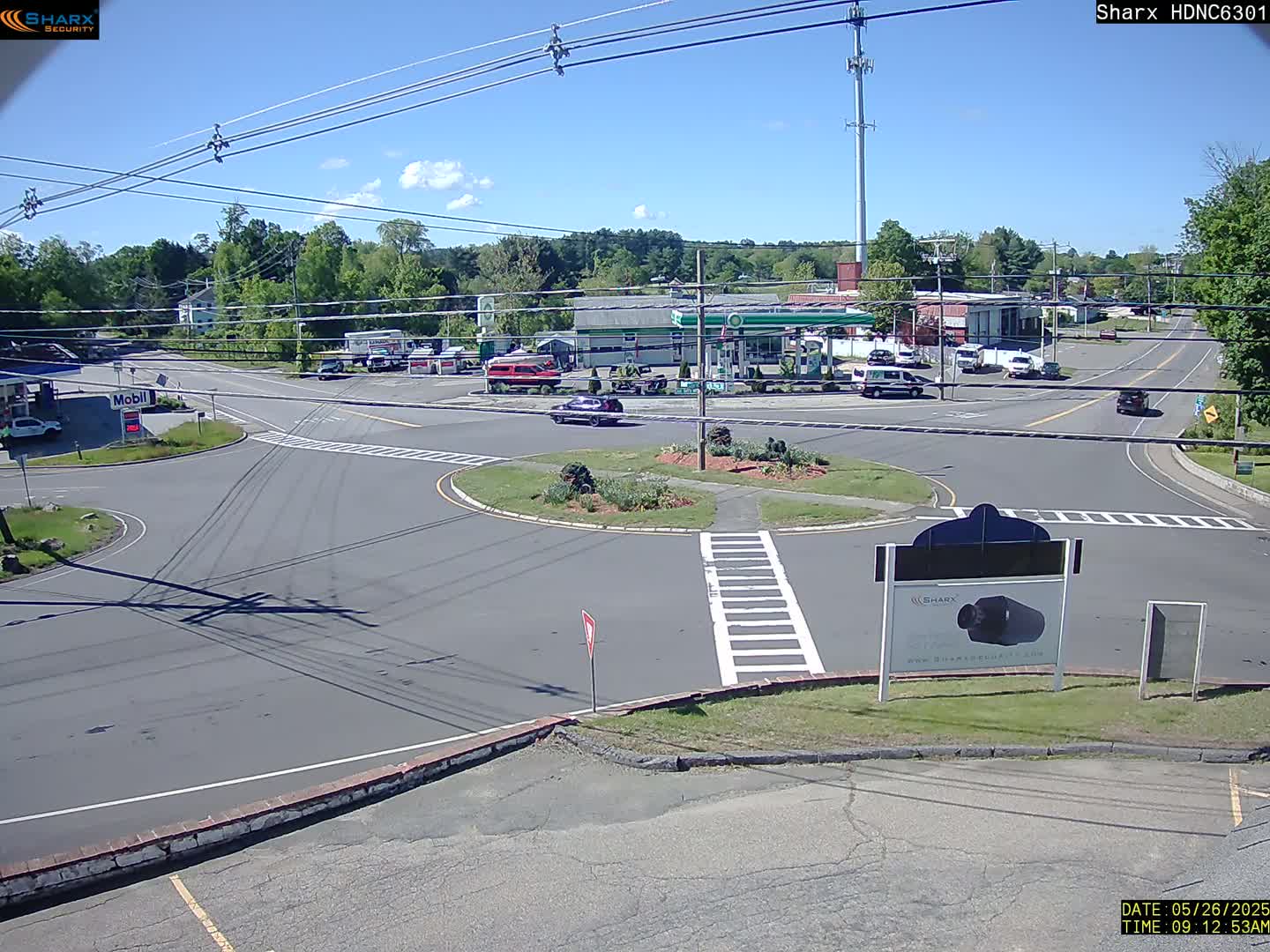 A sunny day shows a view of a roadway intersection with a traffic circle, surrounded by businesses and trees.