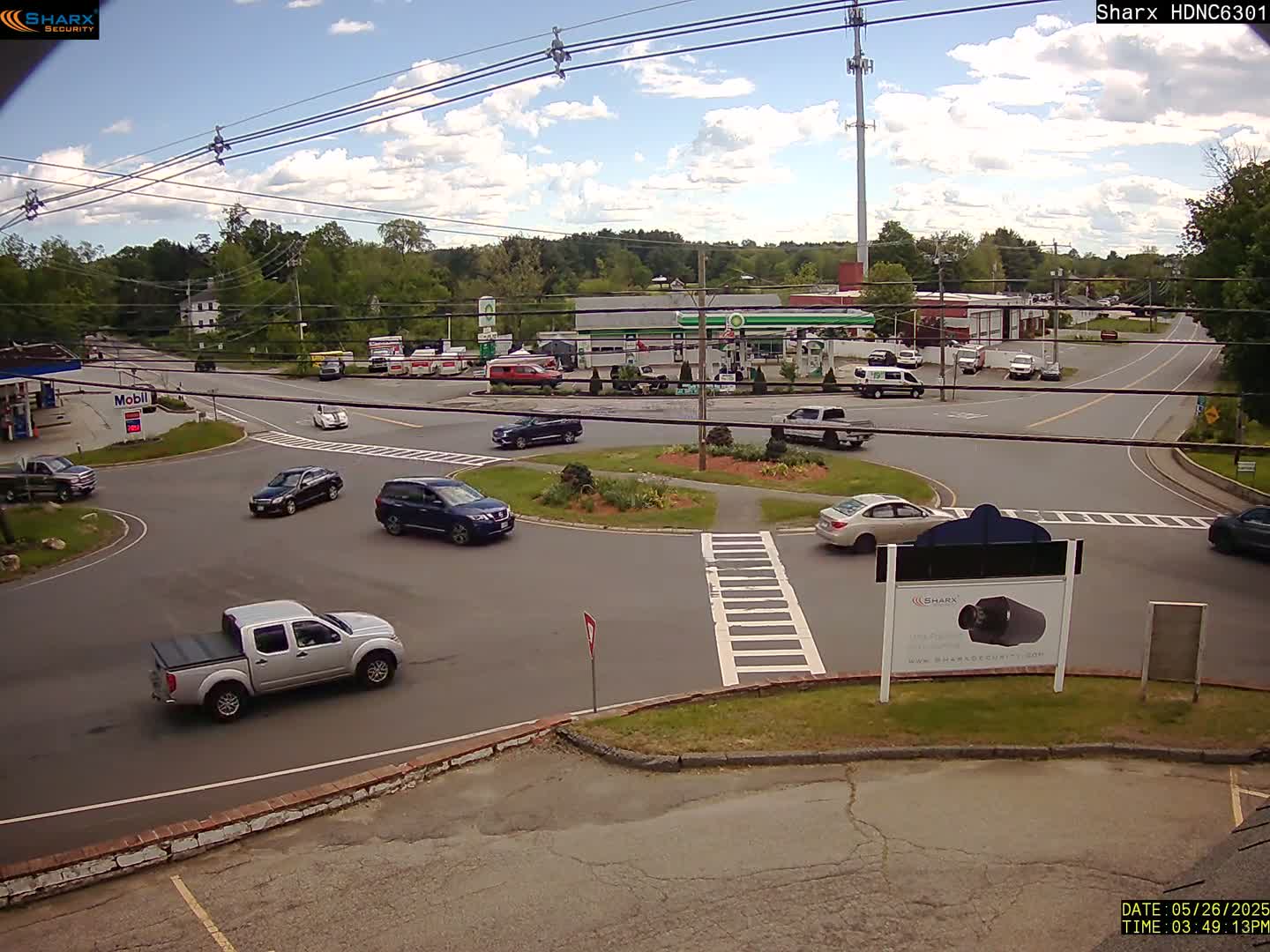 A partly sunny day shows several cars navigating a roundabout and intersection near a gas station and other businesses, with trees and greenery in the background.