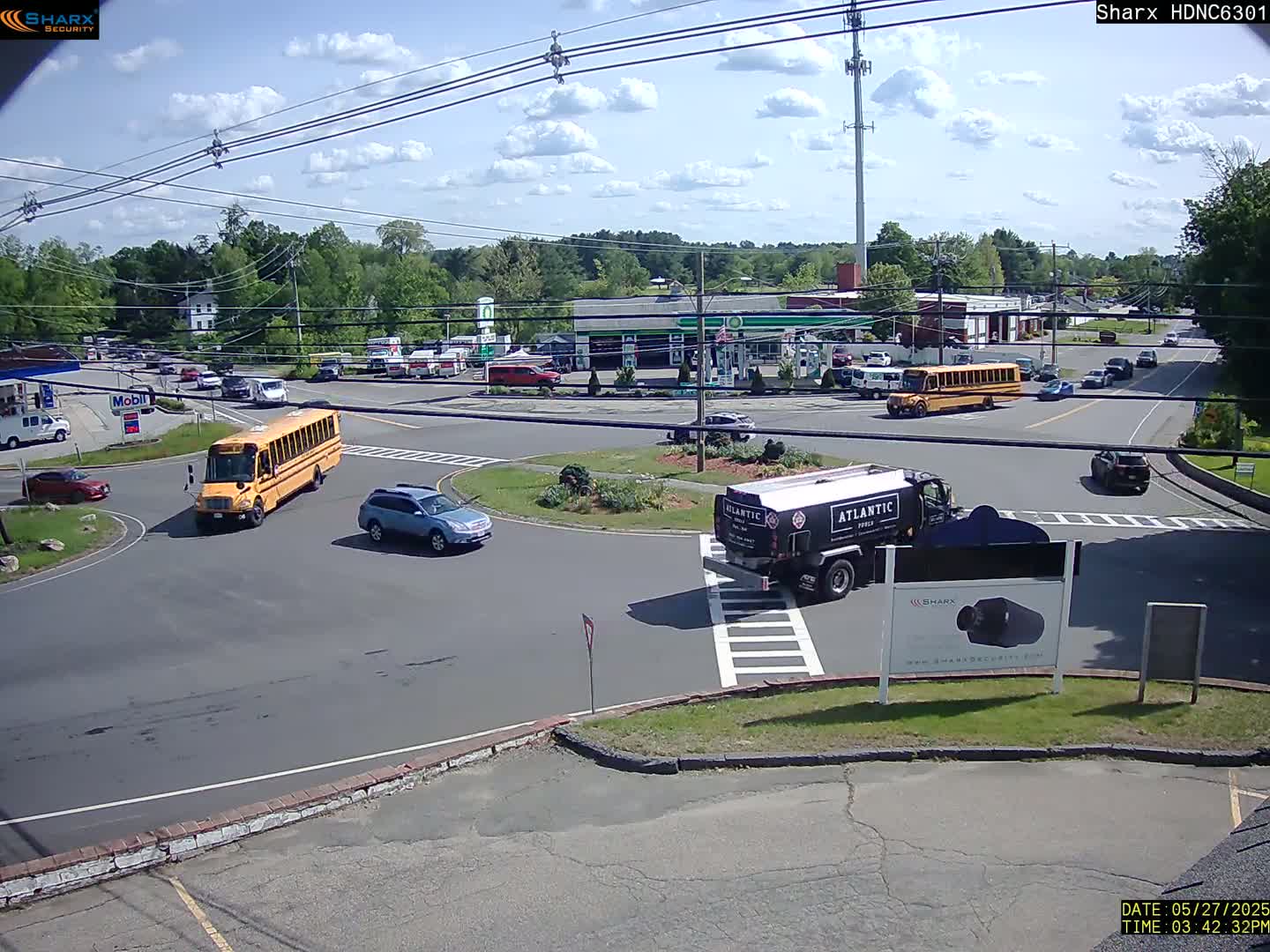 A sunny day shows several vehicles at a crossroads, including two school buses, a fuel tanker, and cars, with a gas station and other buildings visible in the background.