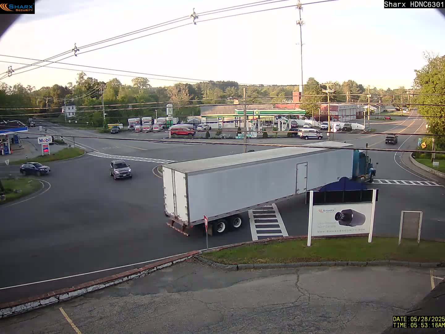 A large white semi-trailer truck is stopped at a crossroads near gas stations and other businesses on a partly sunny day.