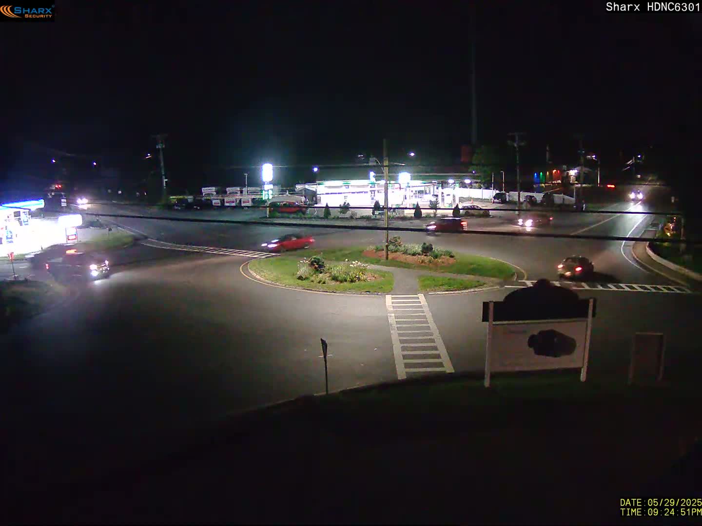 A nighttime view of a roundabout with several cars, a gas station, and surrounding buildings under dark skies.