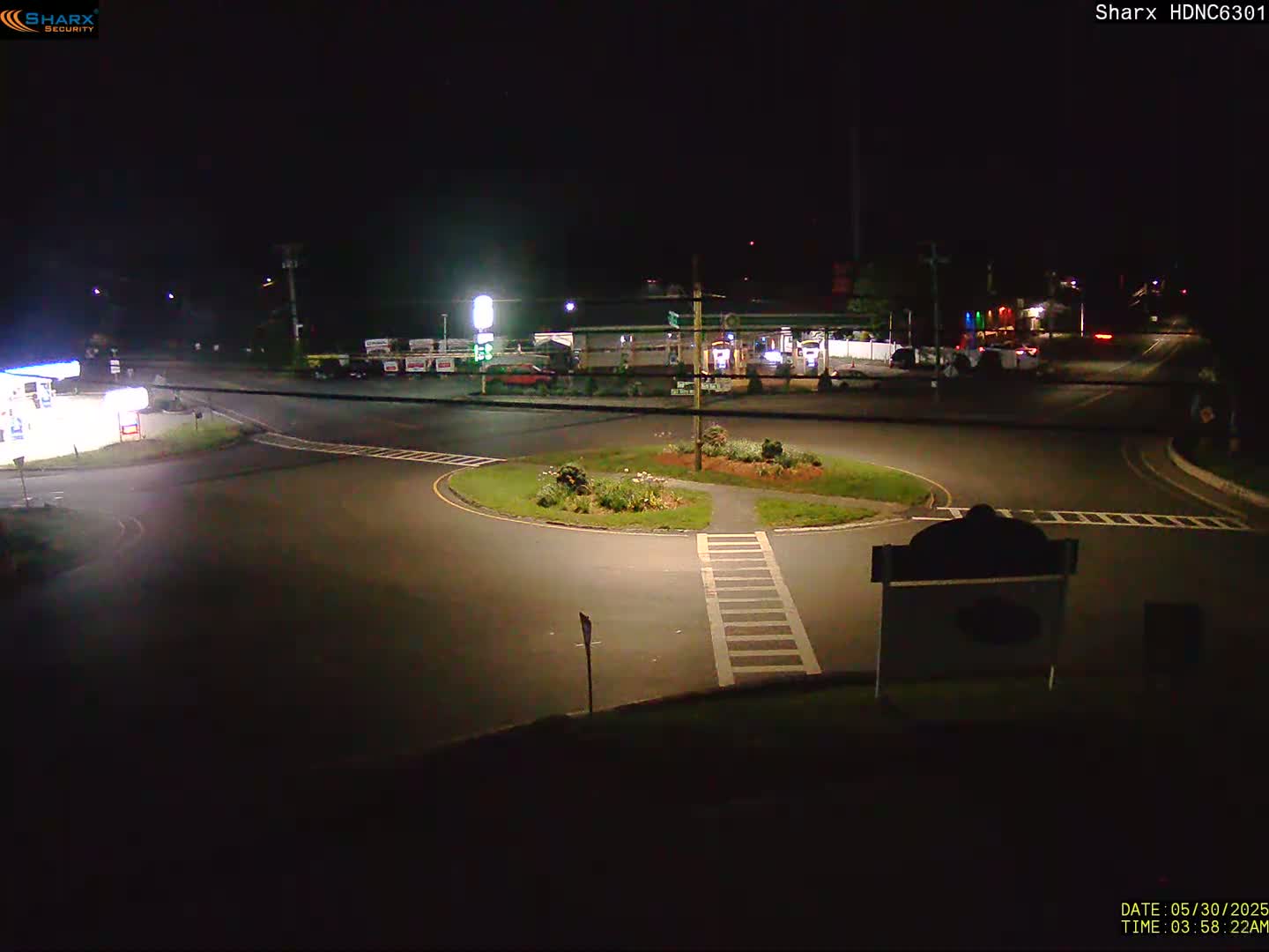 A nighttime view of a roundabout intersection with a gas station and minimal traffic under dark skies.