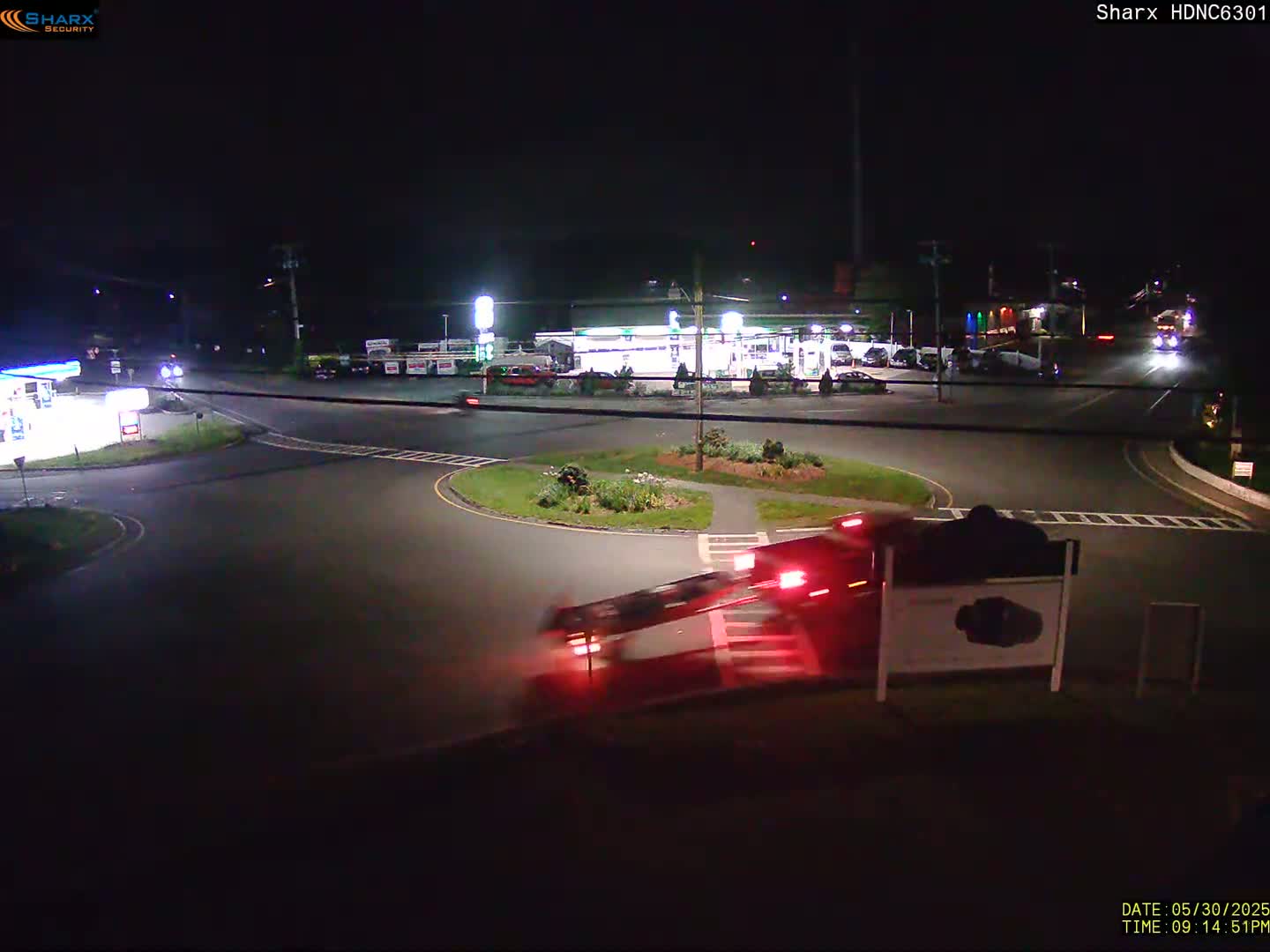 A red vehicle is turning onto a circular intersection at night near a gas station and other businesses.