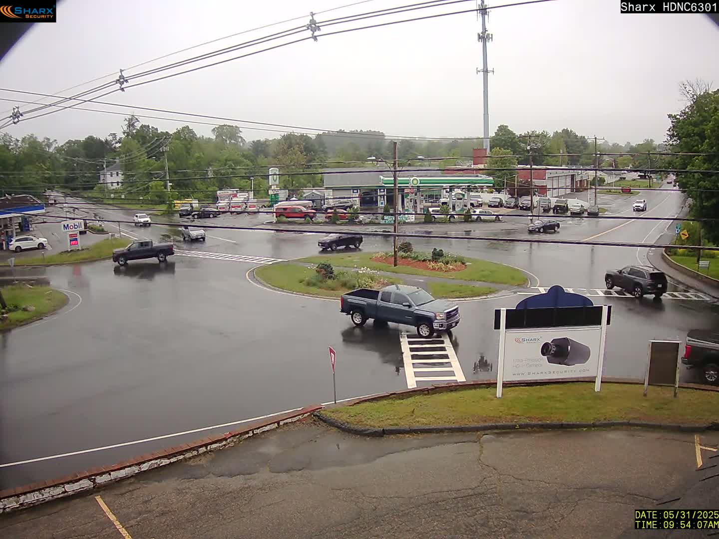 A rainy day scene shows a traffic circle with several vehicles and a gas station in the background, surrounded by trees and buildings.
