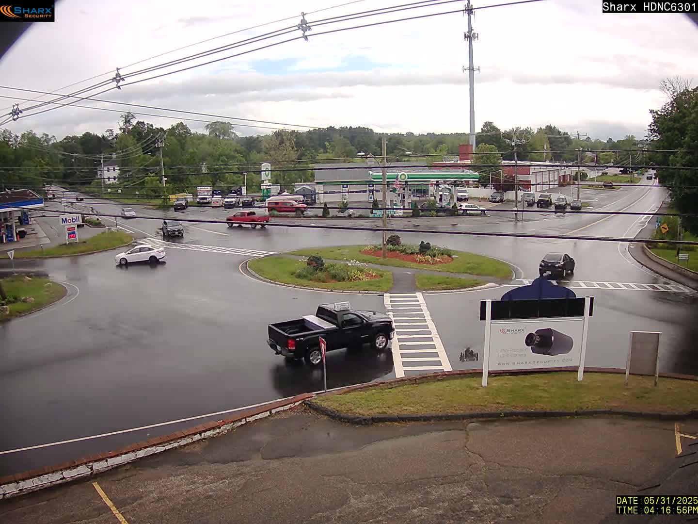 A wet roundabout and surrounding roads with several vehicles, gas stations, and buildings are visible under an overcast sky.