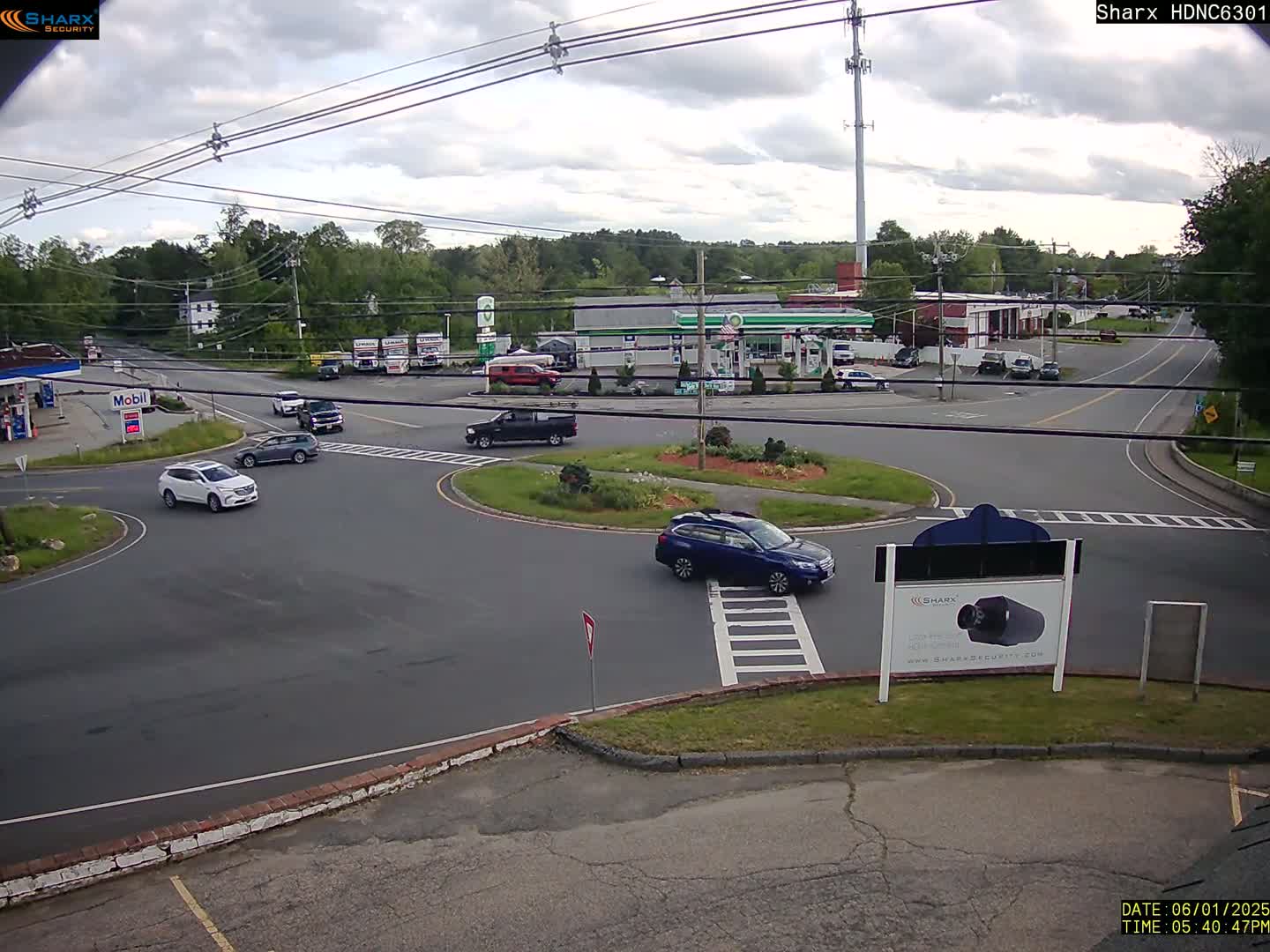 Several cars are driving around a roundabout near gas stations and businesses under a partly cloudy sky.