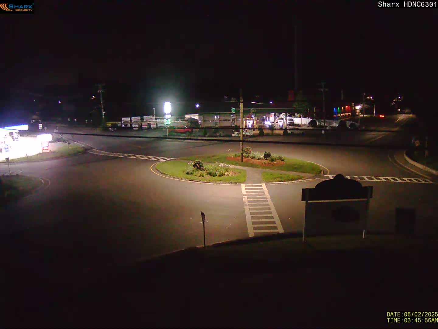 A nighttime view of a circular intersection with a gas station and other businesses visible in the background under dark conditions.