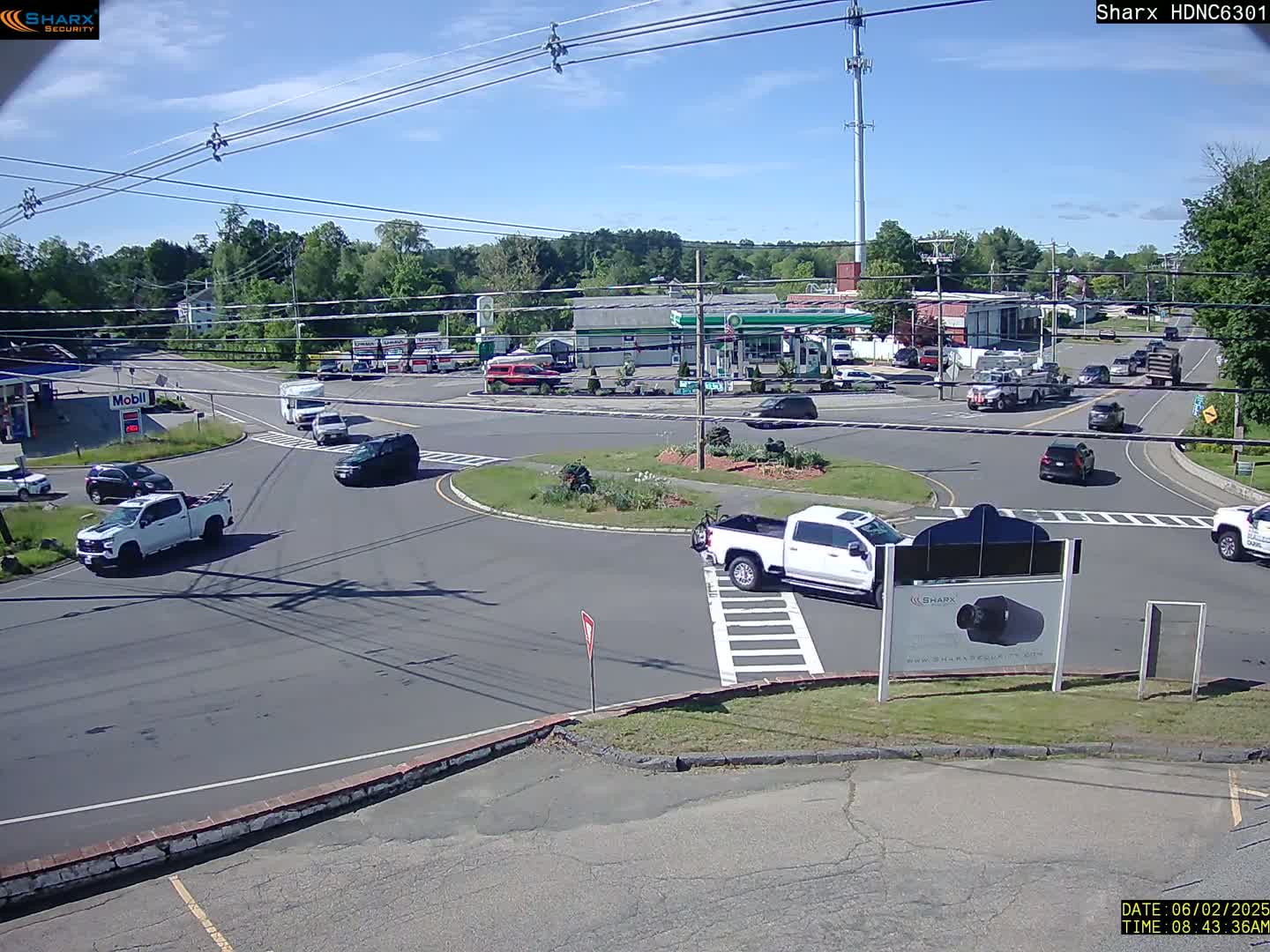 A sunny day shows several vehicles navigating a roundabout near a gas station and other businesses, surrounded by trees.