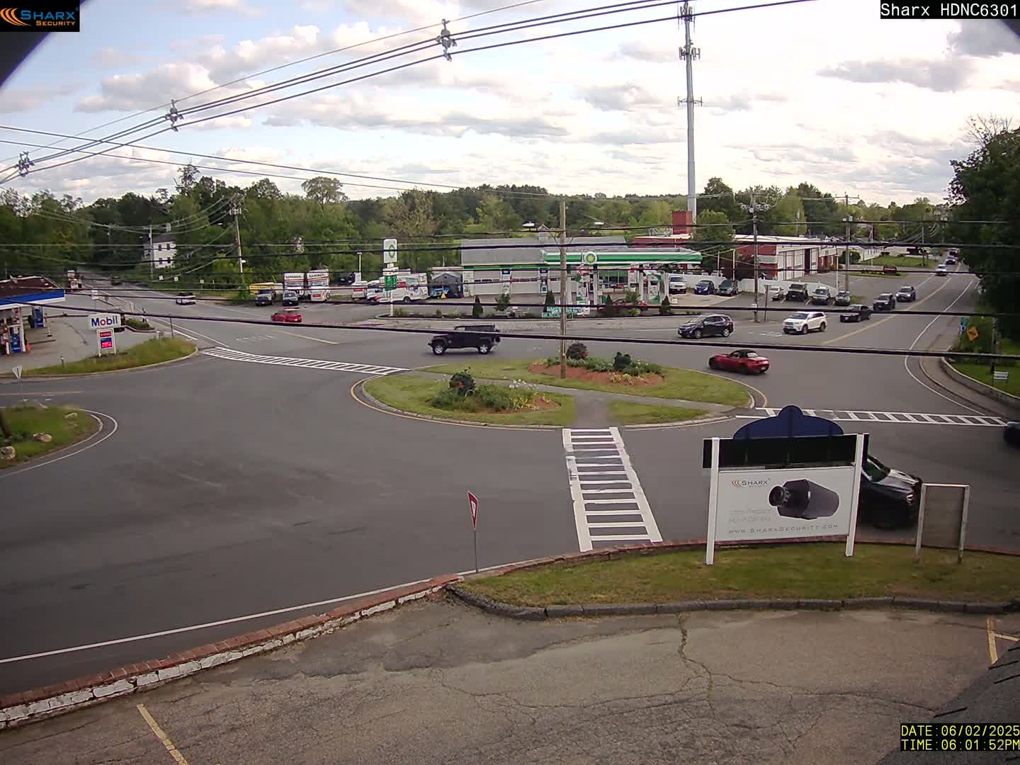 A partly cloudy day shows a roadway intersection with several cars, gas stations, and surrounding businesses.