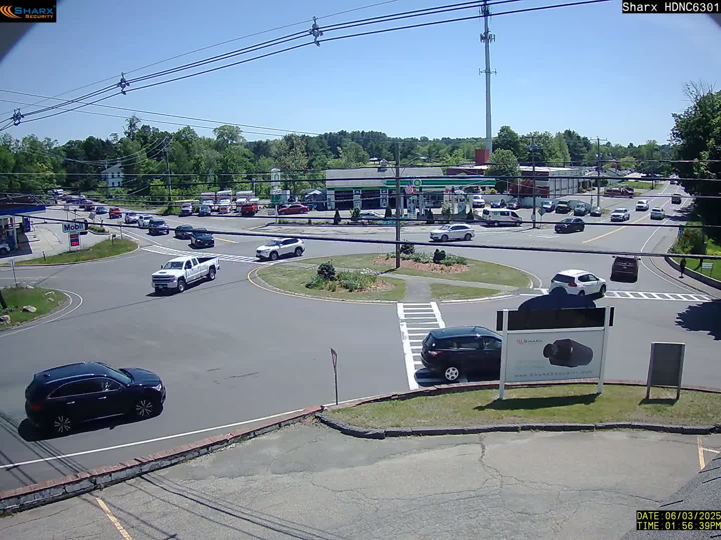 A sunny day shows a roundabout with several cars, a gas station, and surrounding buildings in a suburban area.