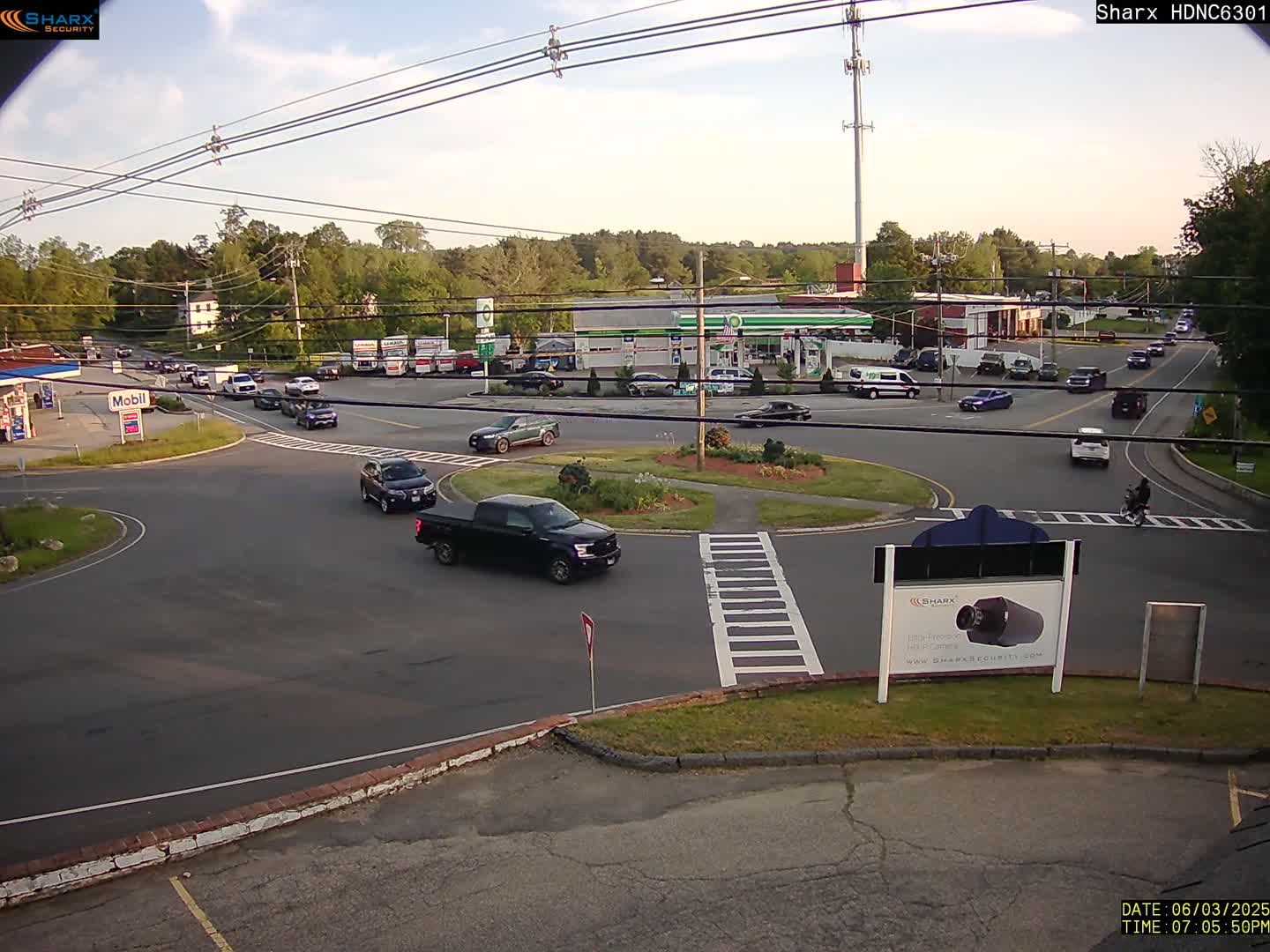 A mostly sunny day shows several vehicles navigating a roundabout near a gas station and other businesses, with trees and greenery in the background.