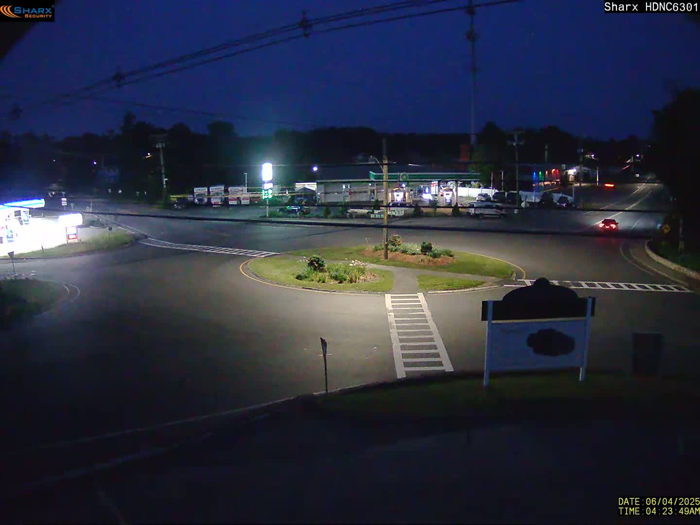 A nighttime view of a roundabout intersection with a gas station and other businesses visible in the background under a dark sky.