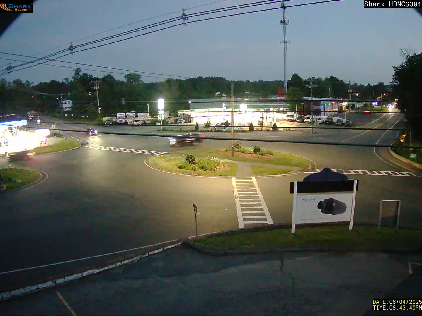 A nighttime view of a roundabout intersection with several cars and a gas station in the background, under a dusky sky.