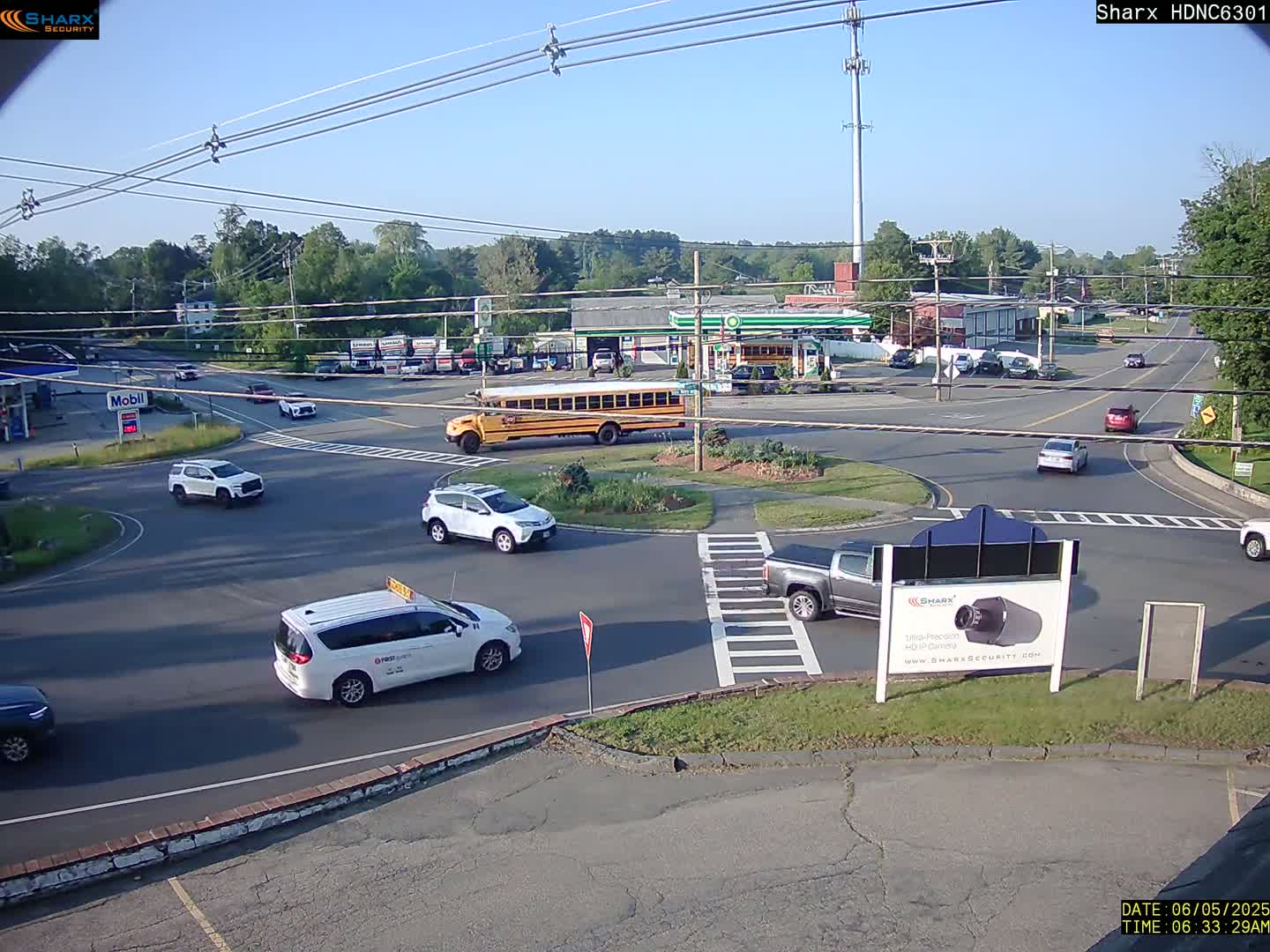 A sunny day shows several vehicles navigating a roundabout near a gas station and school bus, with surrounding greenery and power lines overhead.