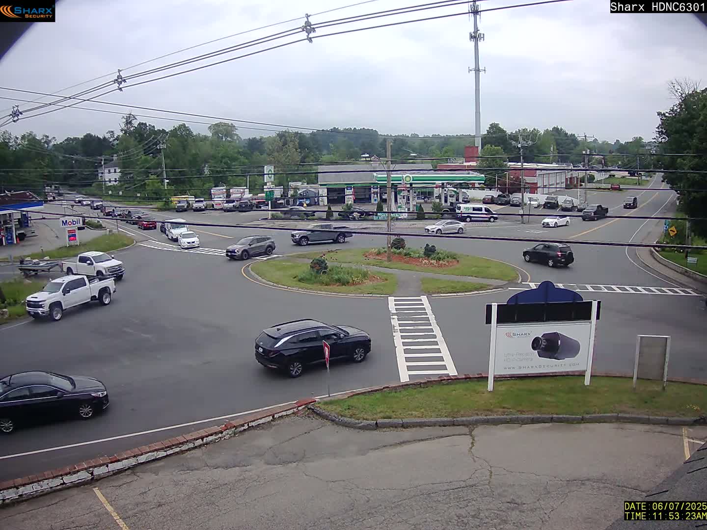A mostly cloudy day shows a roundabout intersection with several cars and trucks, a gas station, and some businesses in the background.