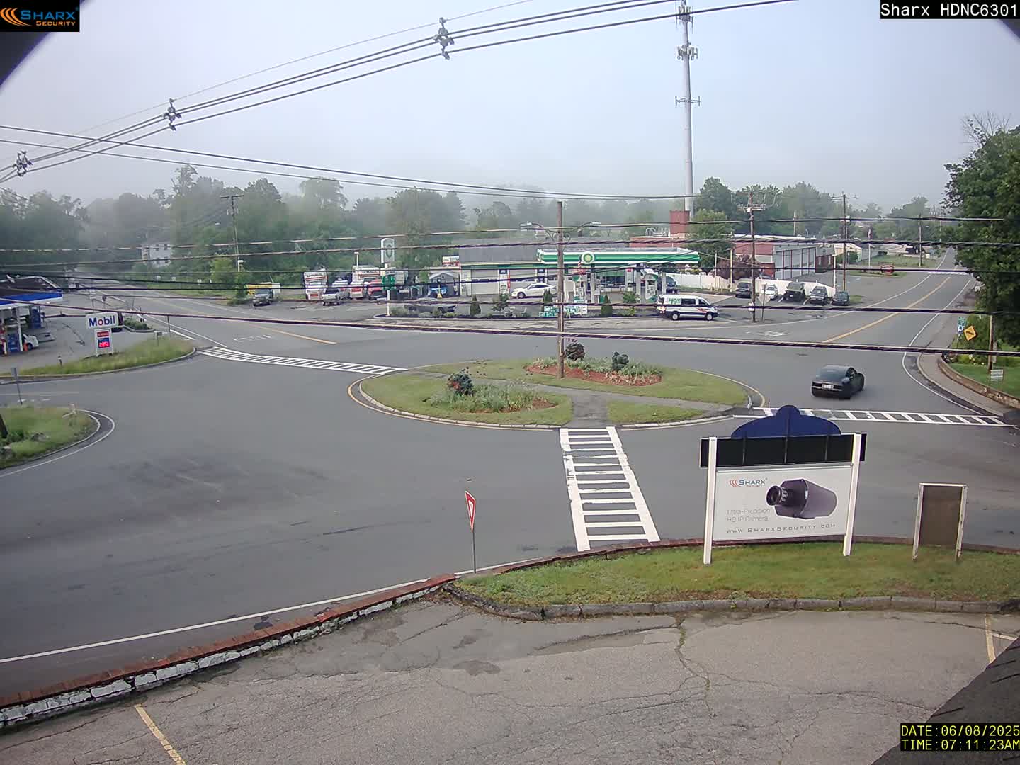 A misty view of a road intersection with a gas station and several cars, centered around a small traffic circle.
