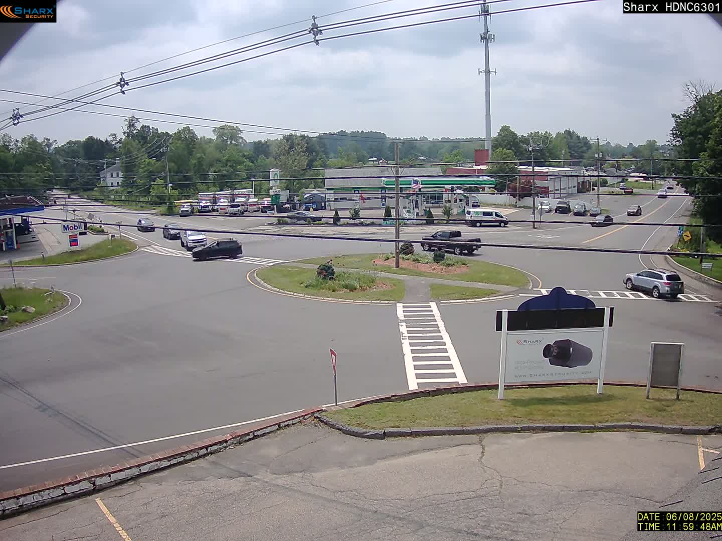 A mostly sunny day shows a multi-lane roundabout with several cars and light traffic, near a gas station and other businesses in a suburban area.