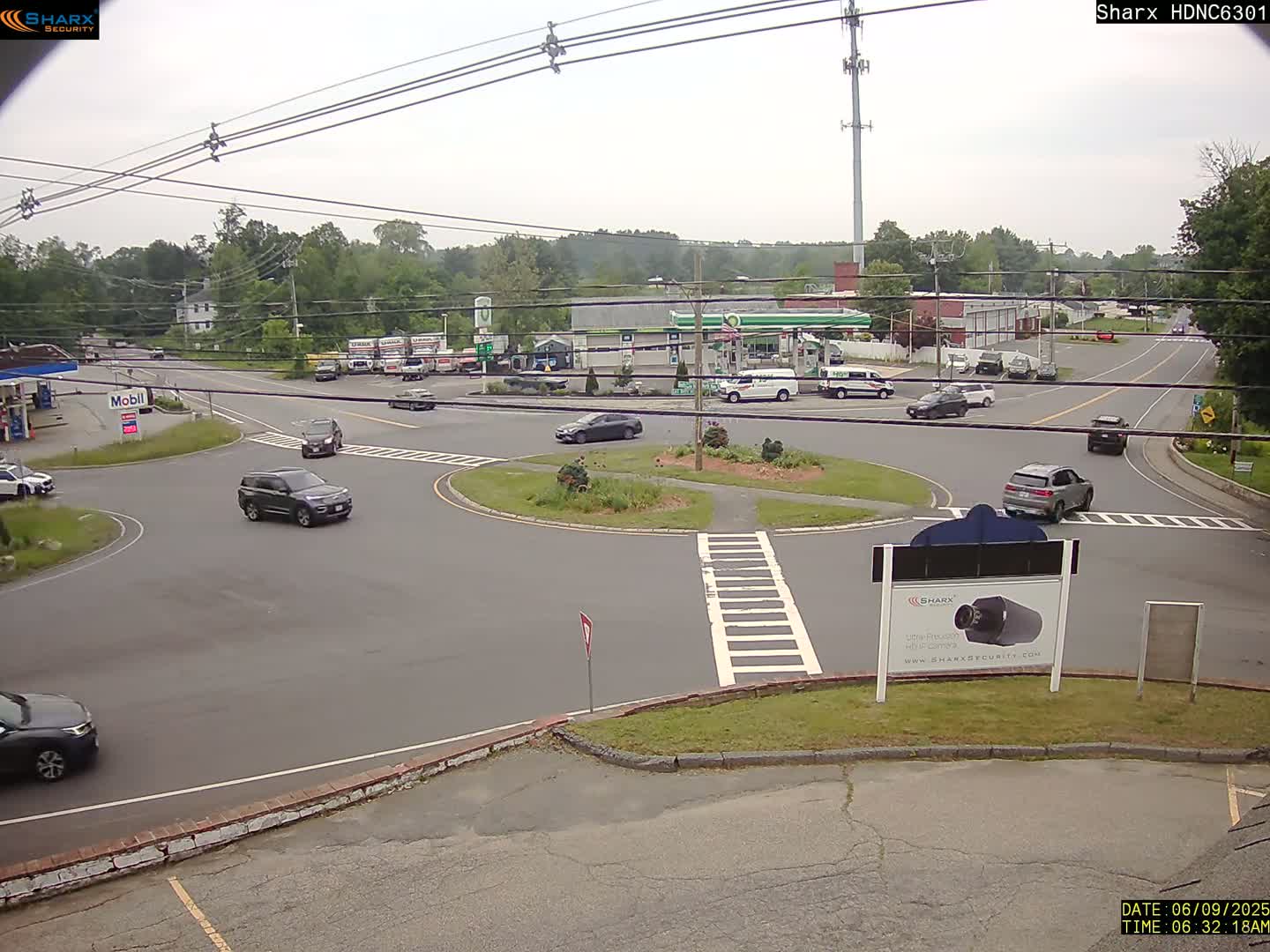 A mostly sunny day shows several vehicles navigating a roundabout near a gas station and other businesses, surrounded by trees and power lines.