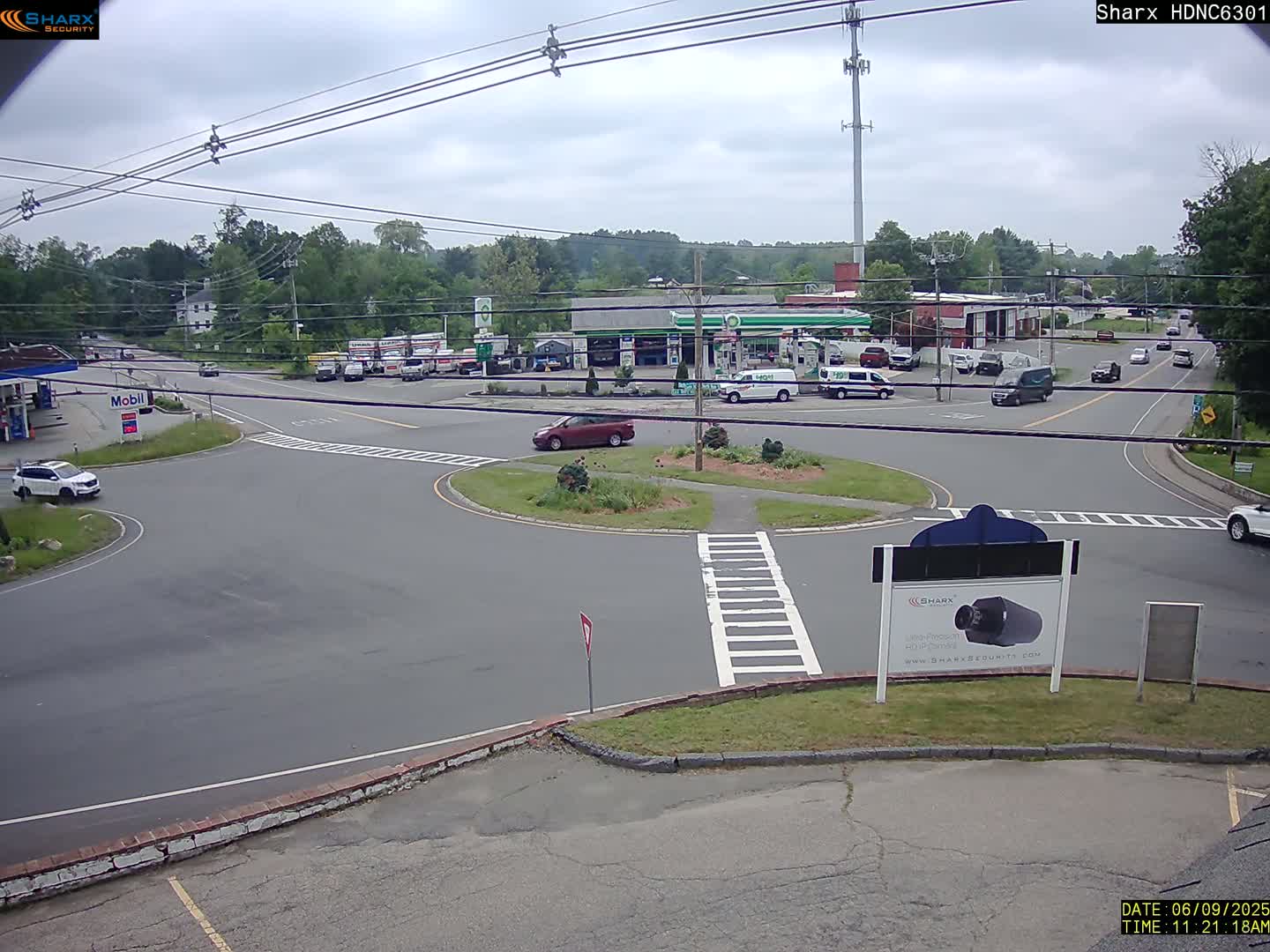 A partly cloudy day shows a traffic circle with several cars and trucks around a gas station and other businesses, with some greenery in the background.