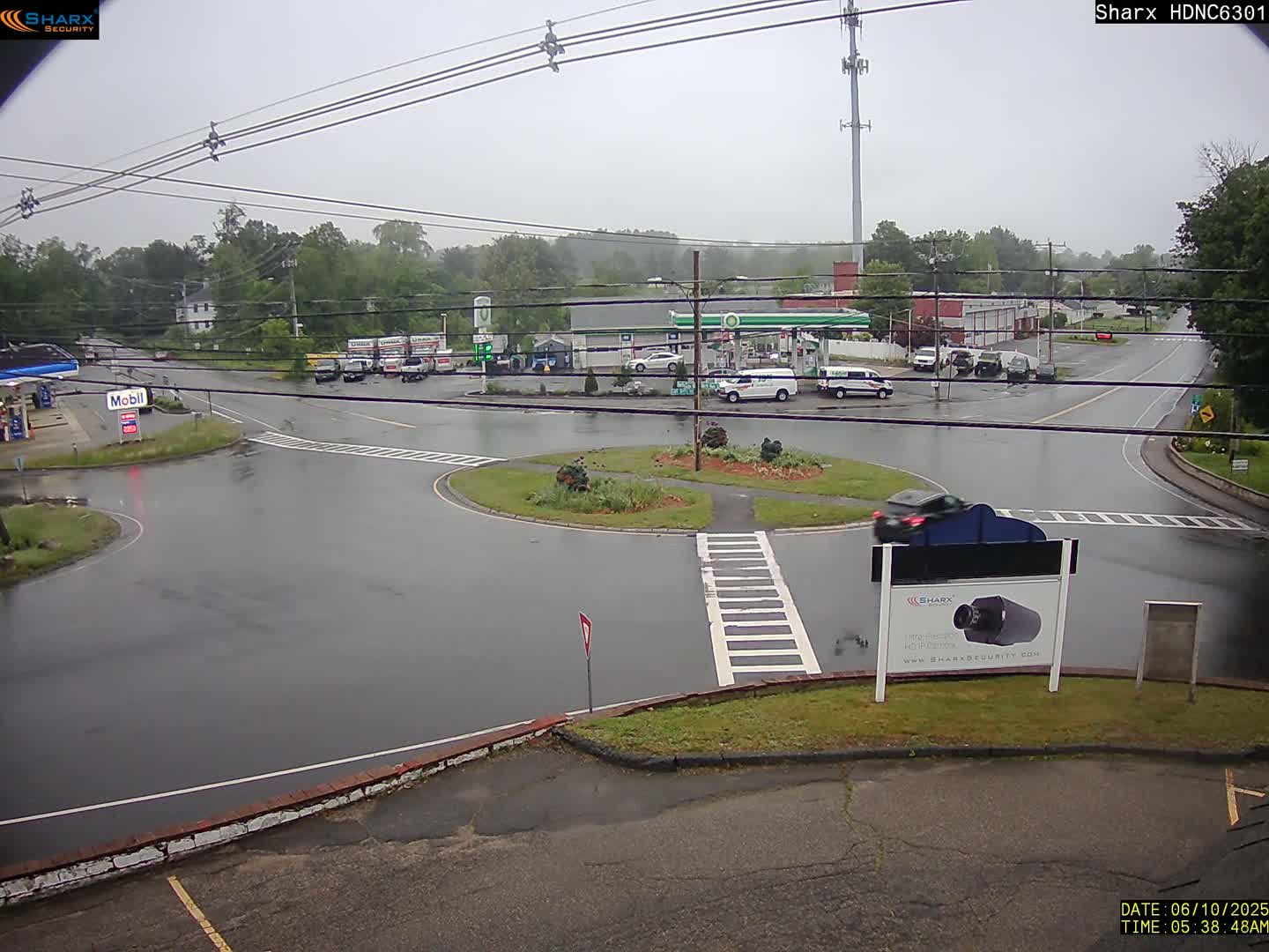 A rainy day at a road intersection shows a gas station and several vehicles, including cars and trucks, in a wet parking lot.