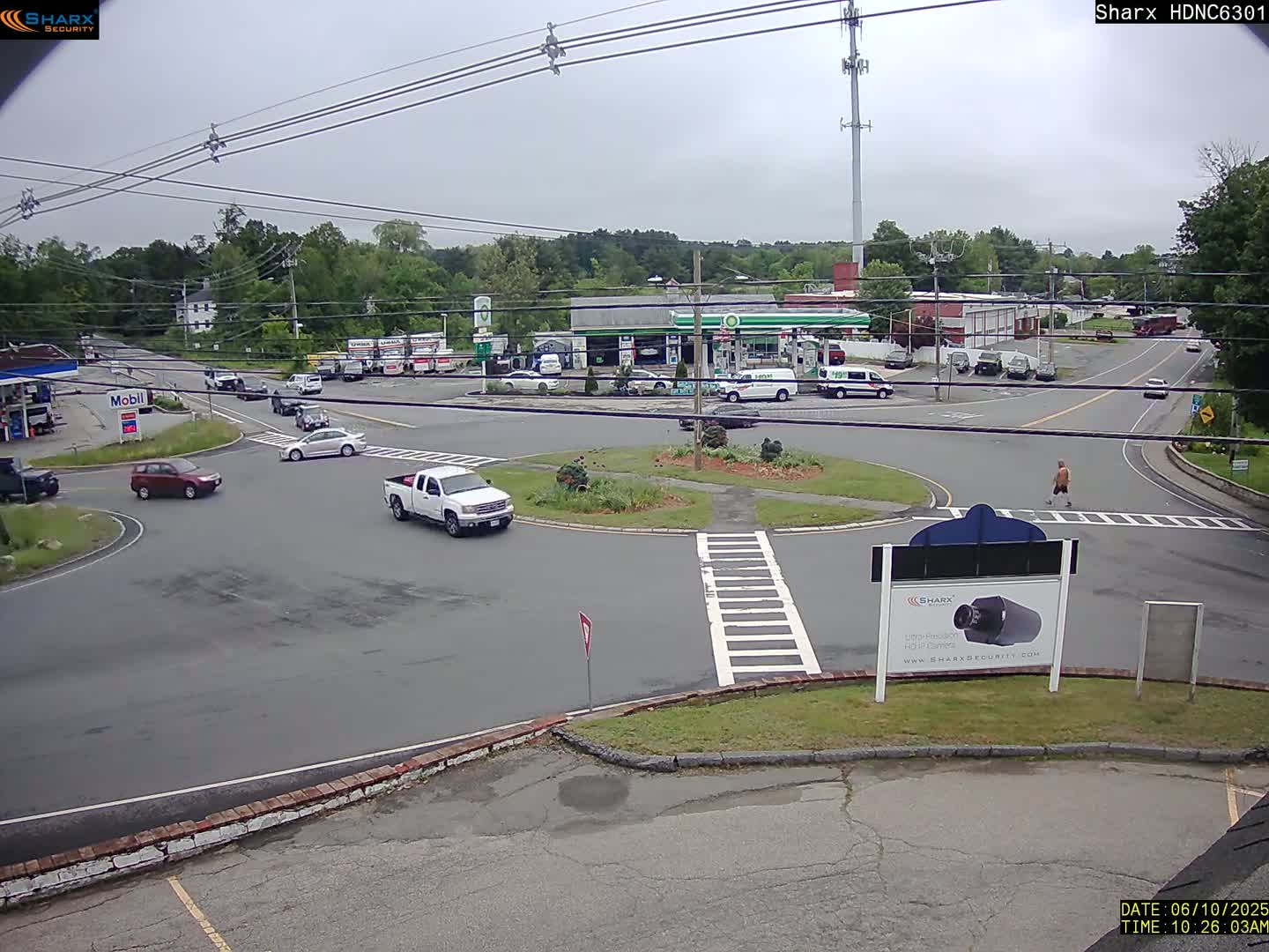 A mostly cloudy day shows several vehicles at a roundabout near a gas station and other businesses, with one pedestrian walking nearby.