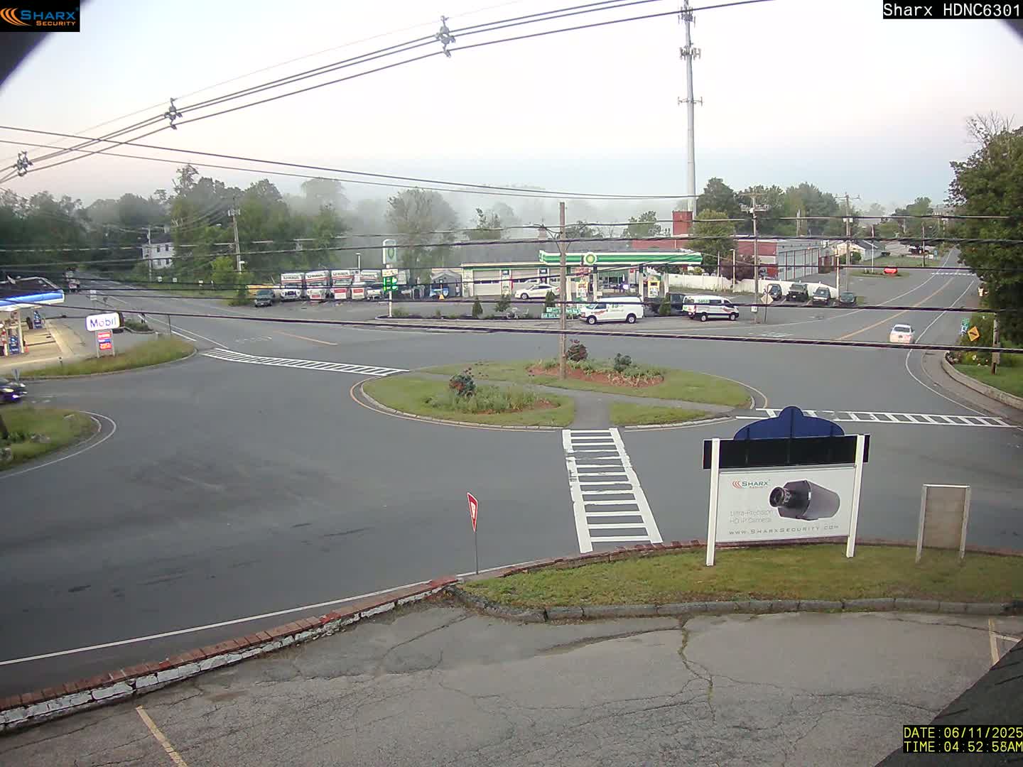 A misty morning scene shows a roundabout intersection with several vehicles and buildings, including a gas station, in the background.