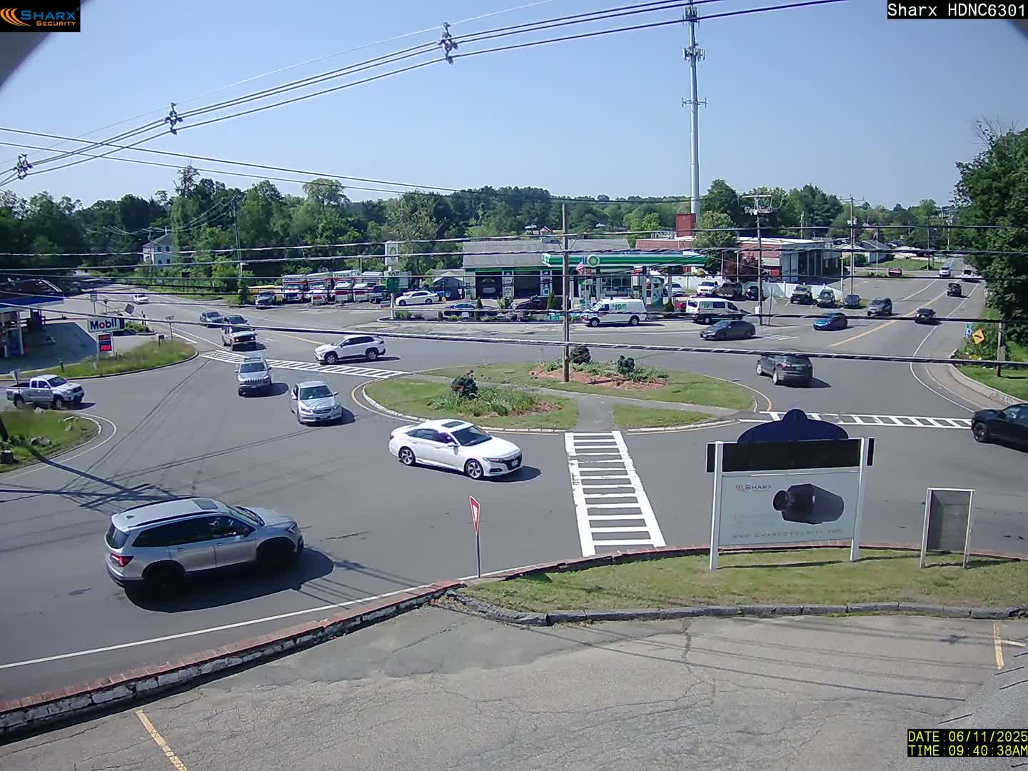 Several cars navigate a roundabout in front of a gas station on a sunny day.