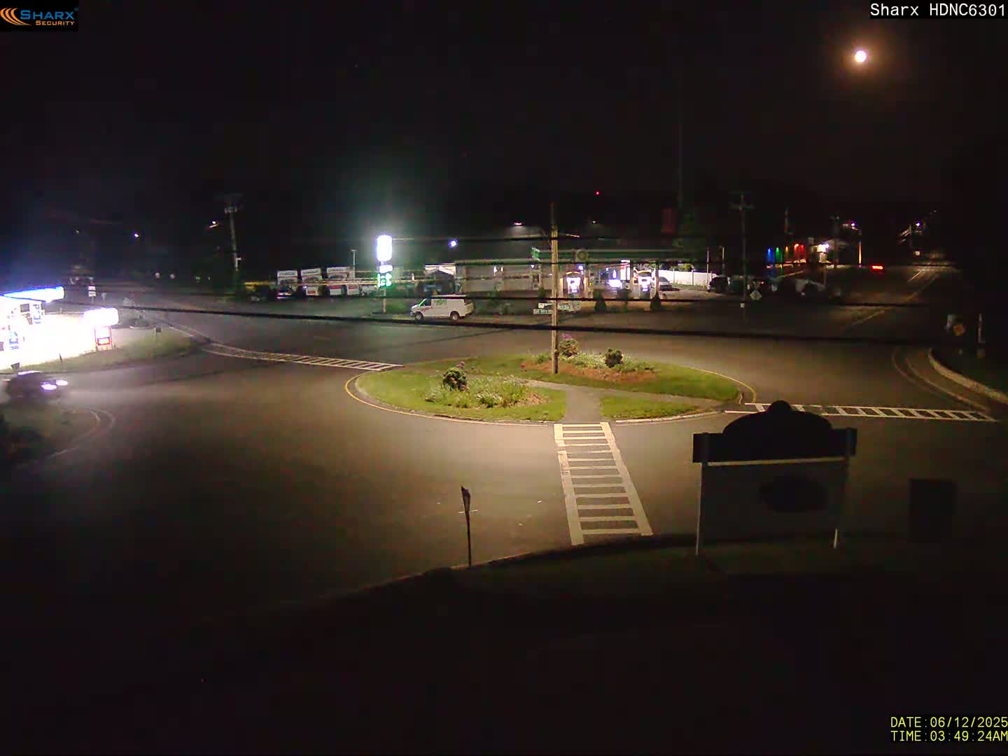 A nighttime view of a gas station and roundabout with a few vehicles, illuminated by streetlights under a bright moon.