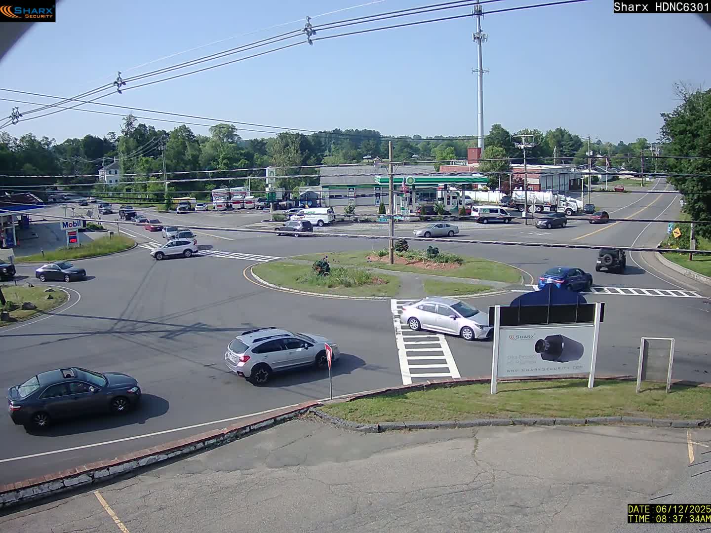 Several cars are driving around a traffic circle near a gas station on a sunny day.