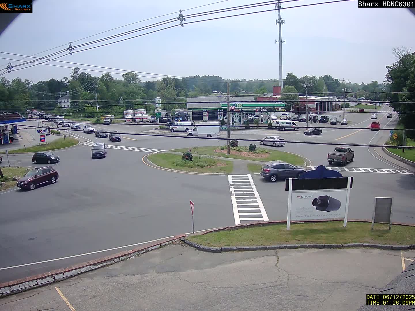 A mostly sunny day reveals a roundabout intersection with several cars and trucks, gas stations, and businesses in the background.