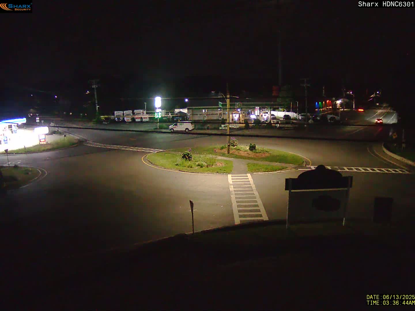 A nighttime view of a mostly empty roundabout intersection near a gas station and other businesses under dark skies.