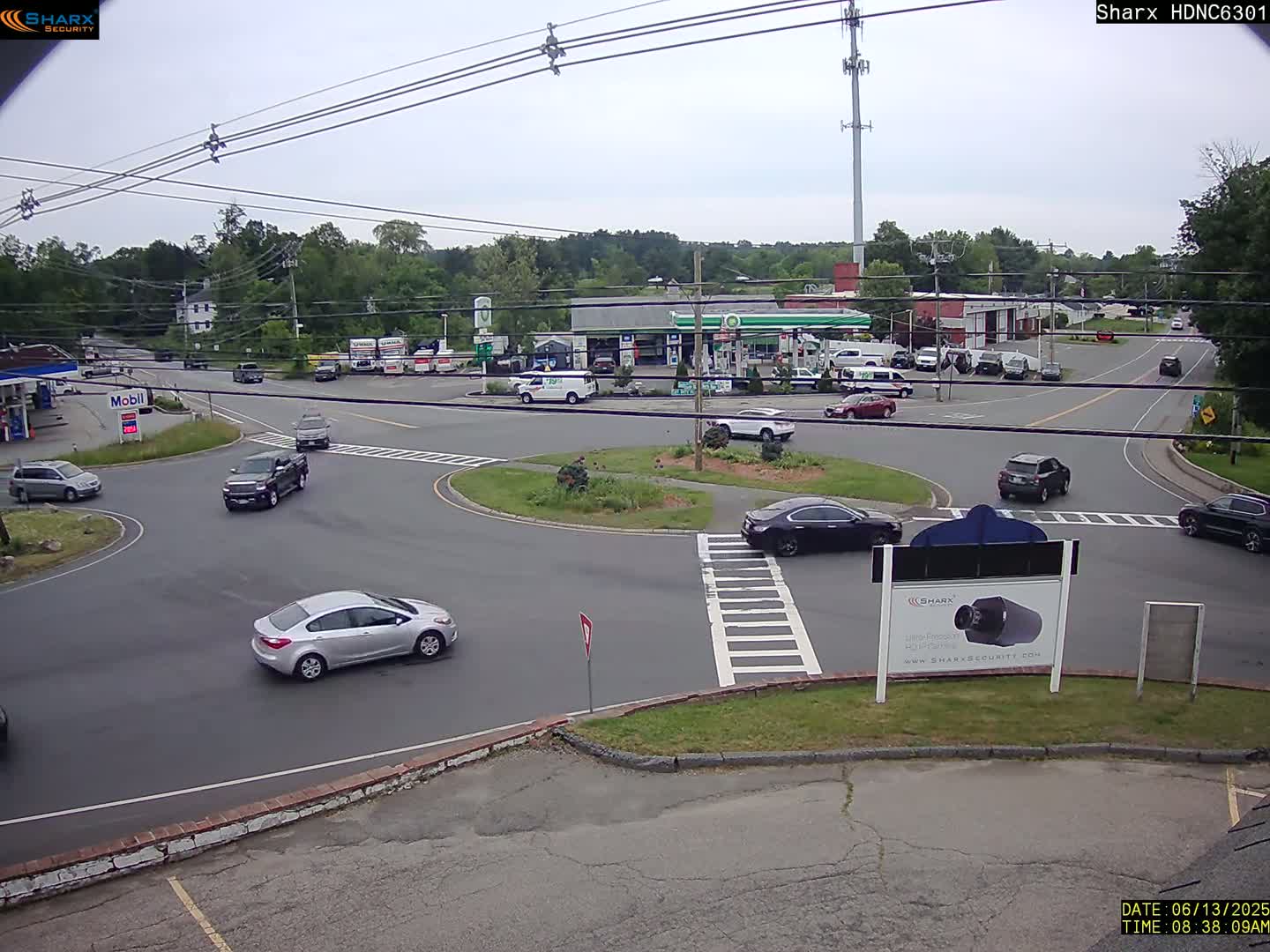 Several cars are driving around a traffic circle near a gas station and other businesses on an overcast day.