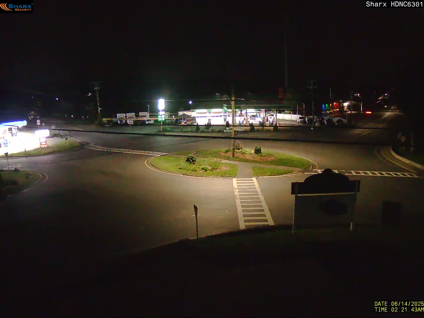 A sunny day shows a traffic circle with several cars and a motorcycle, surrounded by businesses and green trees.