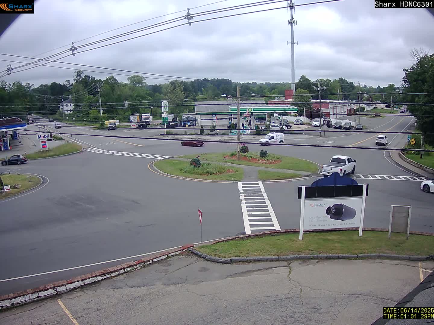 A cloudy day shows a view of a roundabout intersection with several cars, gas stations, and businesses, surrounded by trees.