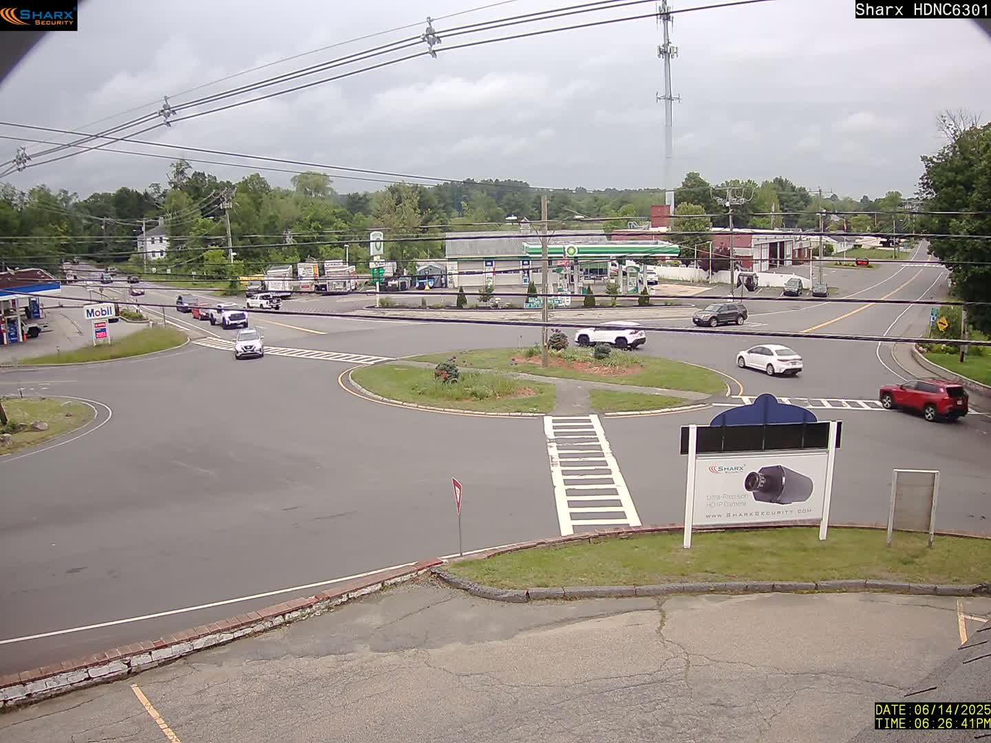 A cloudy day shows a view of a roundabout intersection with several cars, gas stations, and businesses, surrounded by trees.