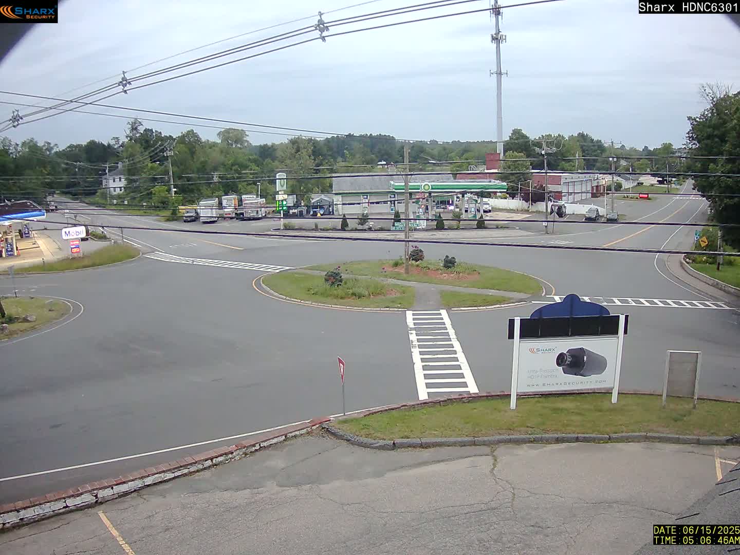 A mostly sunny day reveals a roadway intersection with a gas station and surrounding businesses, all situated amidst green trees.