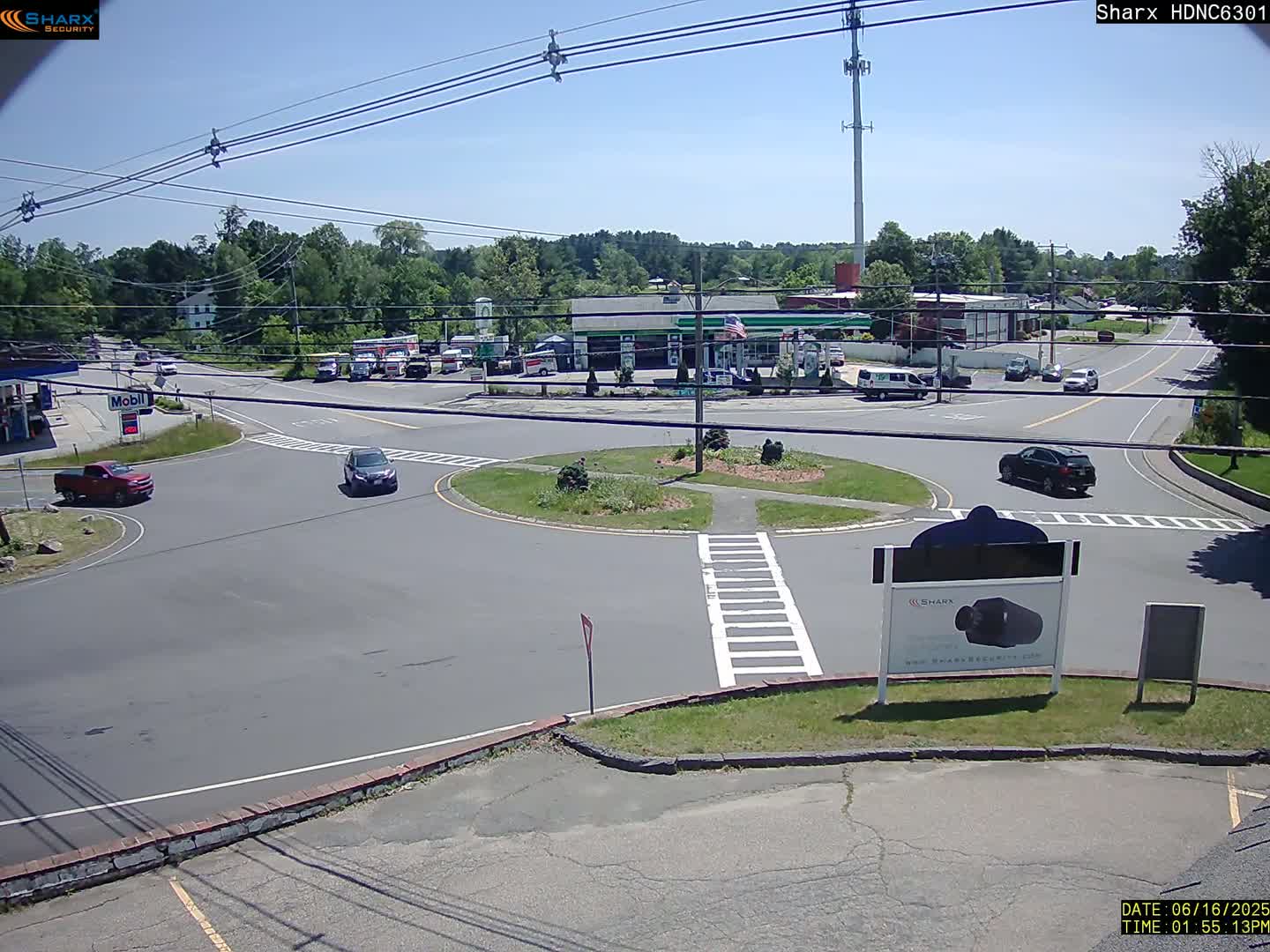A sunny day reveals a traffic circle with several cars, a gas station, and surrounding buildings in a suburban area.