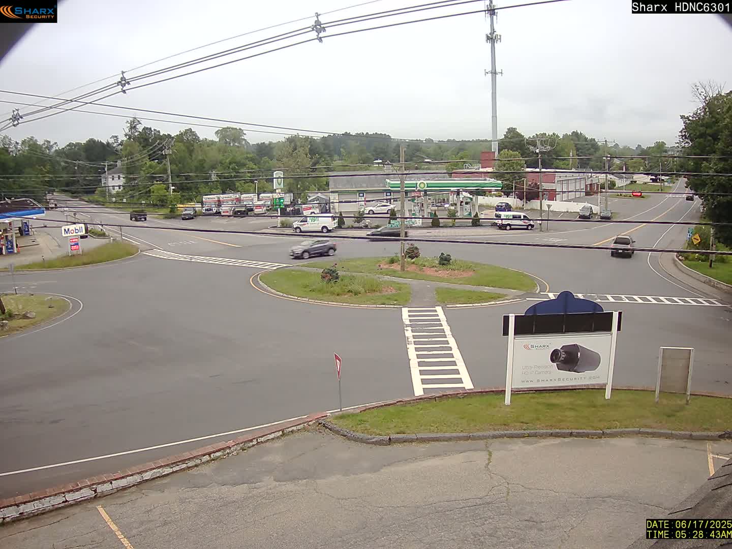 A mostly sunny day reveals a roundabout intersection with several vehicles, a gas station, and surrounding businesses, all set amidst a backdrop of trees.