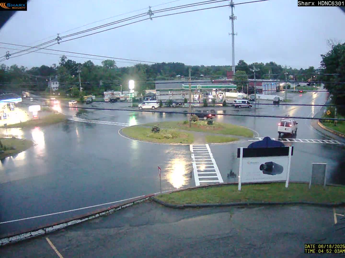 A wet, overcast roadway scene shows a roundabout with several vehicles, including vans and cars, near a gas station and other businesses.