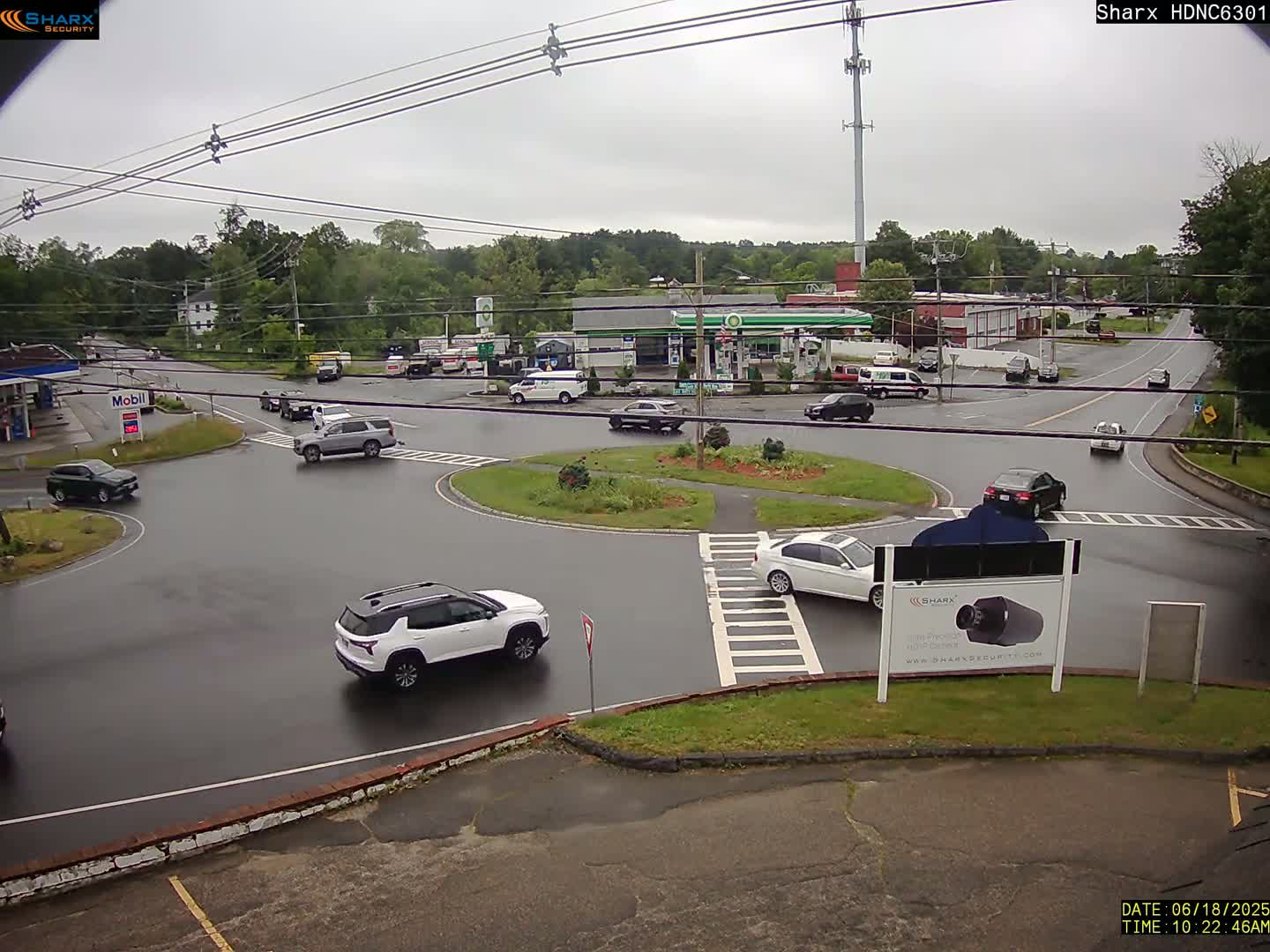 A wet roundabout with several cars and a gas station in the background under an overcast sky.