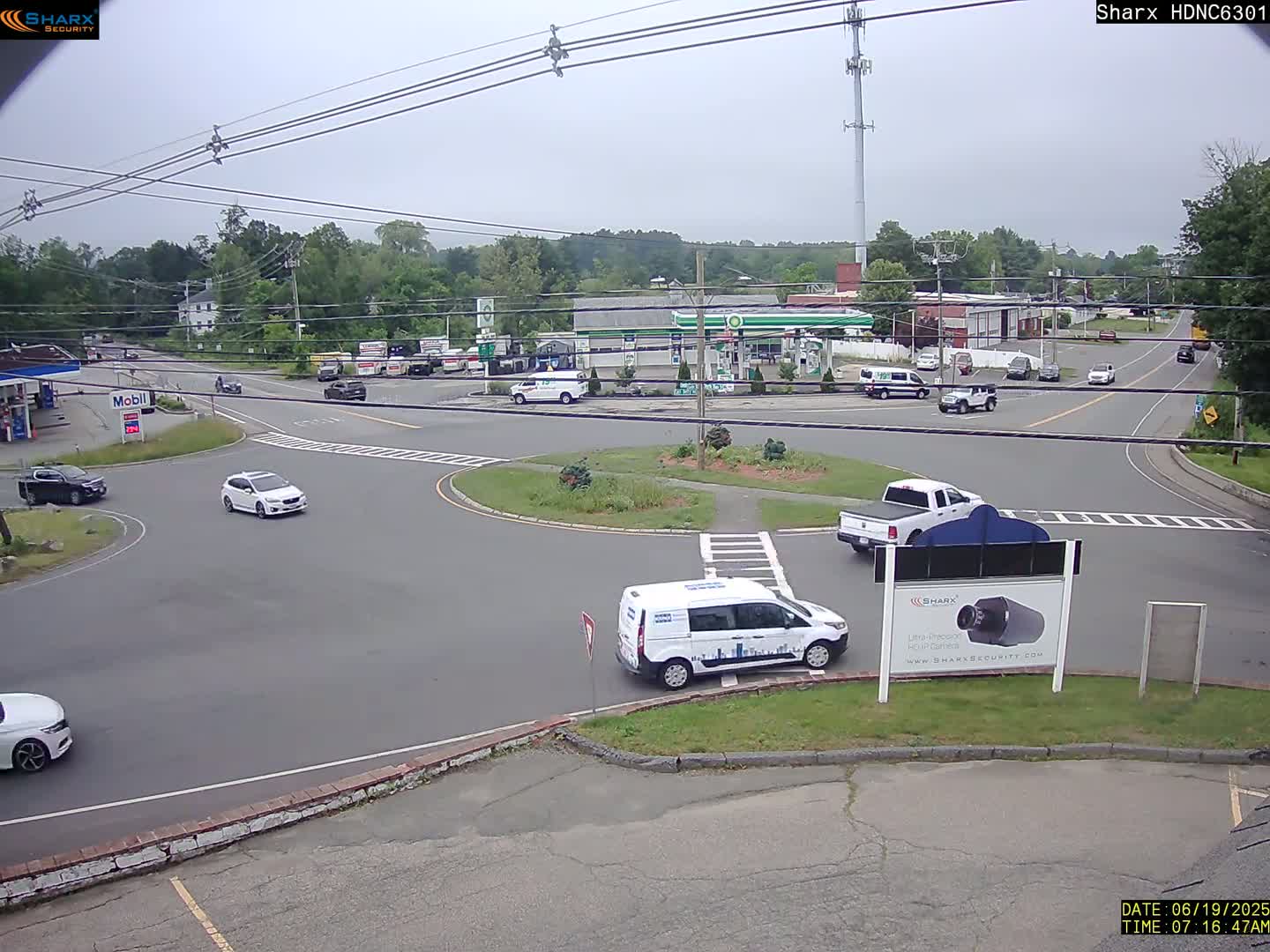 A mostly sunny day shows a roundabout intersection with several cars and vans, near a gas station and other businesses, surrounded by green trees.