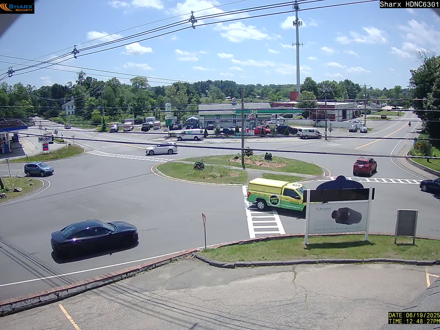 A sunny day shows a roundabout intersection with several cars and light commercial vehicles, near gas stations and other businesses, surrounded by trees.