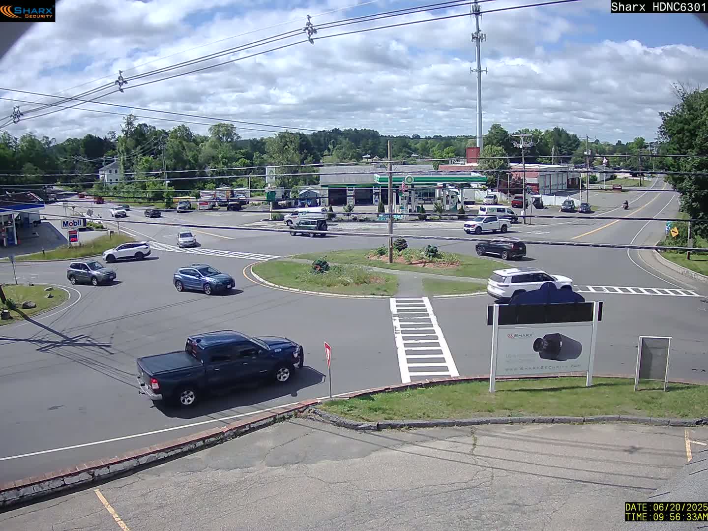 Several vehicles are driving around a traffic circle on a partly cloudy day.
