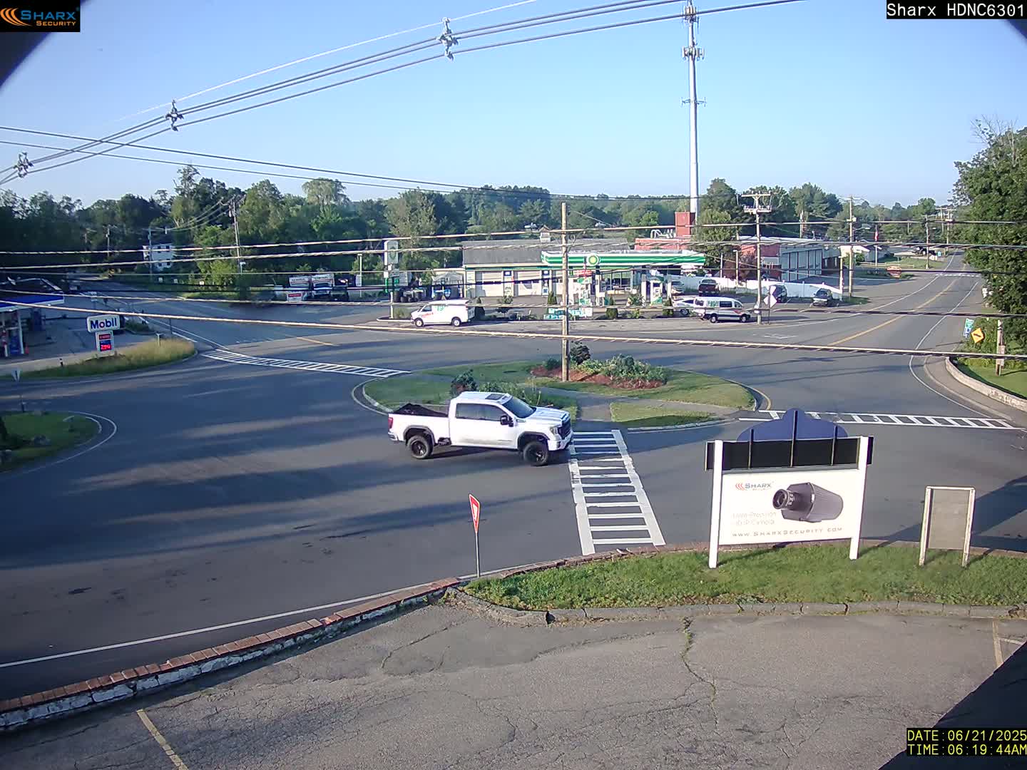 A white pickup truck is driving through a roundabout near a gas station and other businesses on a sunny morning.